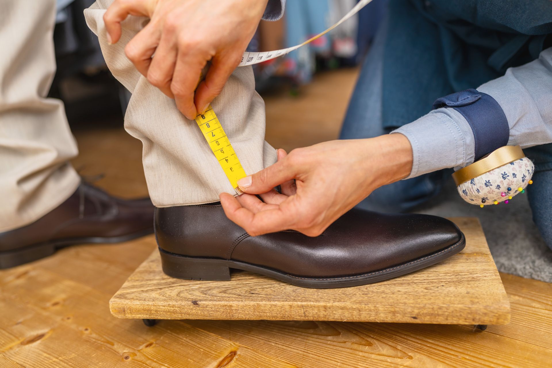 Hands measuring a brown dress shoe with a yellow tape measure on a wooden block