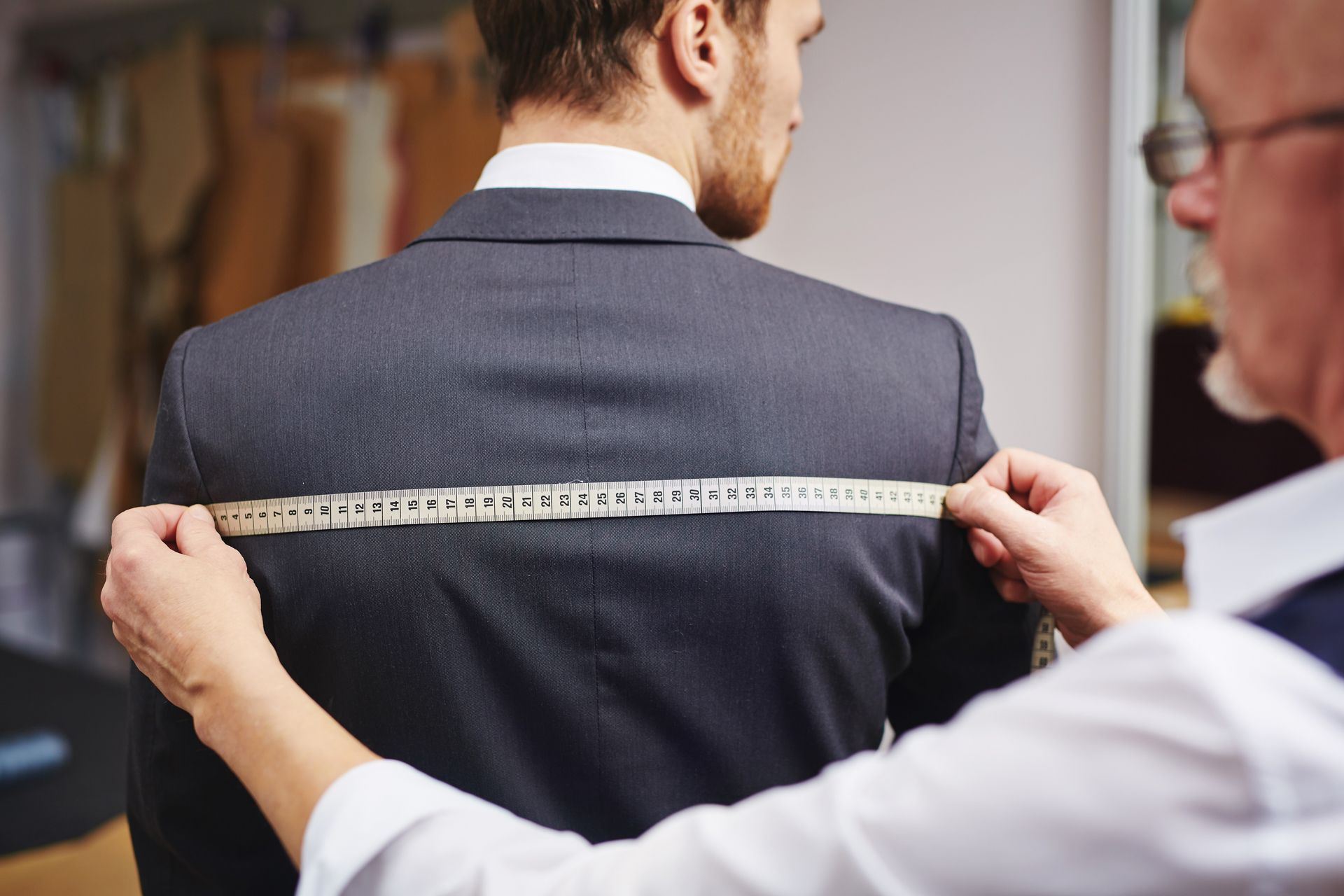 Tailor measuring a man’s suit jacket with a tape measure from shoulder to shoulder