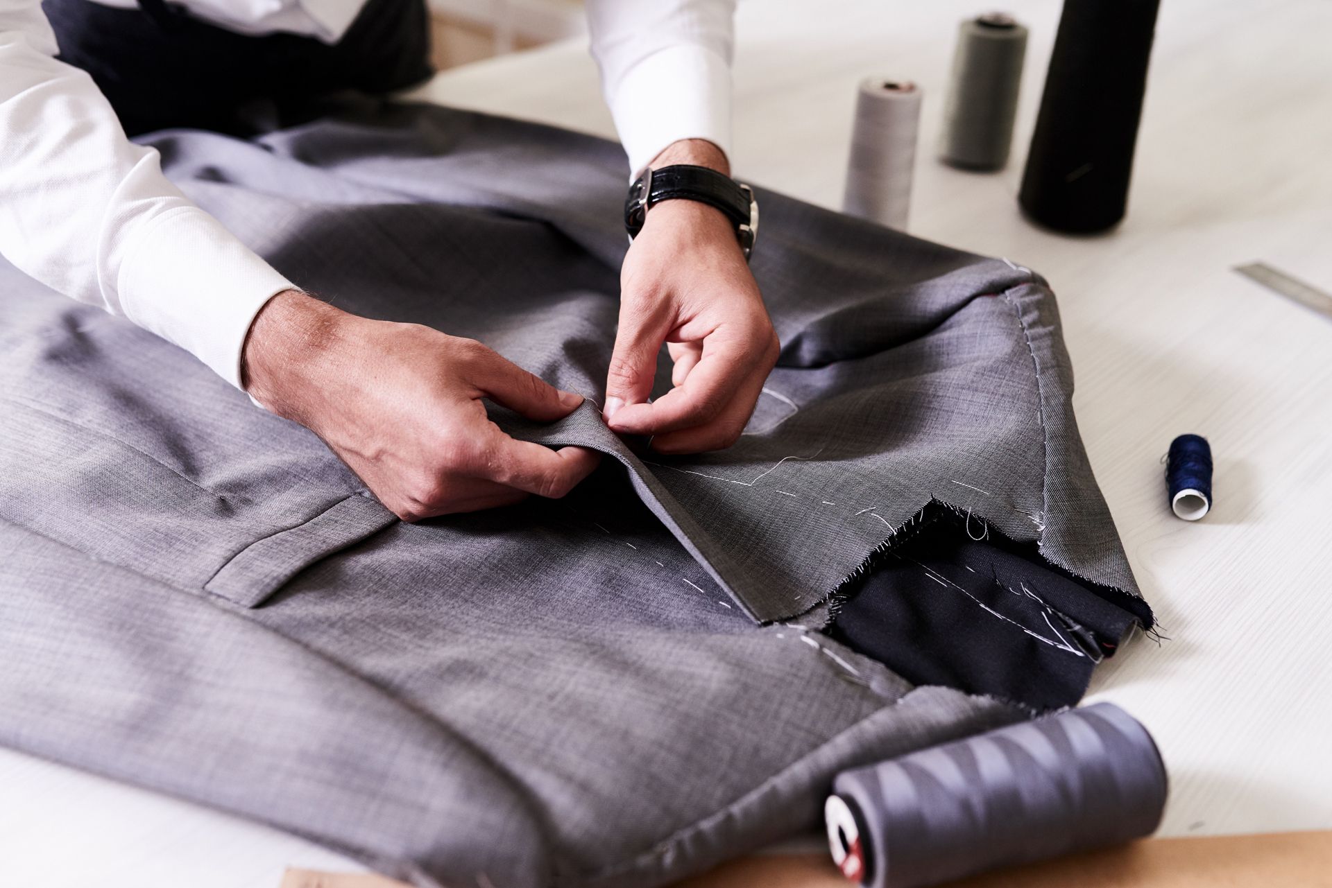Hands pinning gray fabric while sewing tools and thread spools sit nearby on a worktable