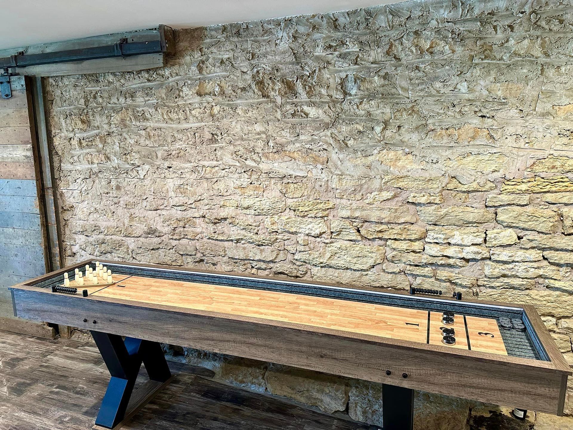Shuffleboard table with wooden surface and dark frame against stone wall.