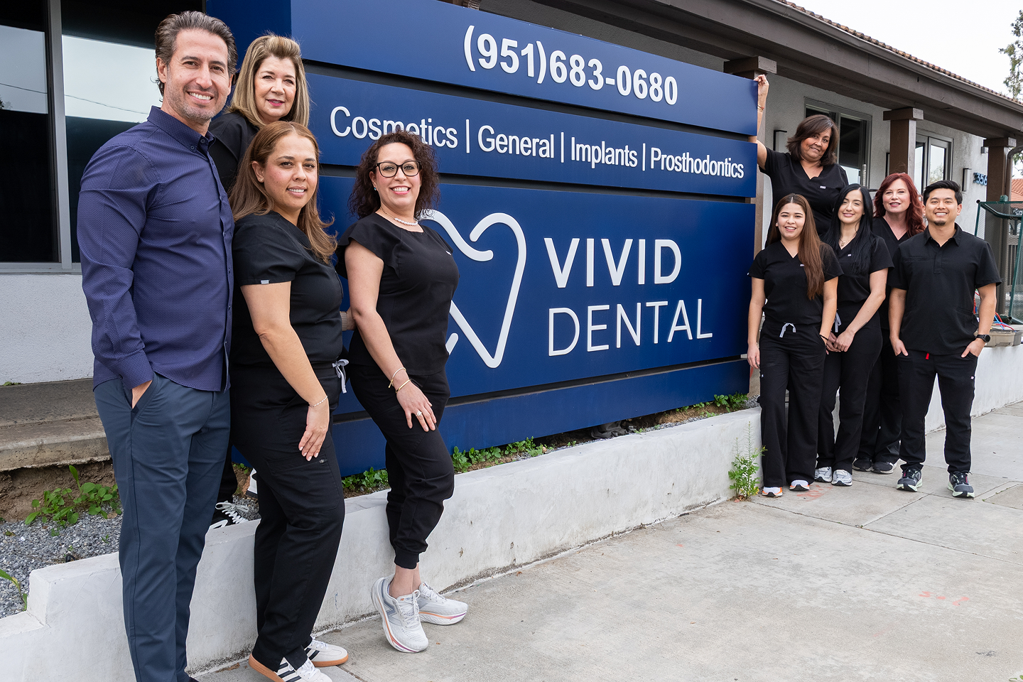 Group of dental staff posing outside Vivid Dental clinic with a blue sign and smiling faces