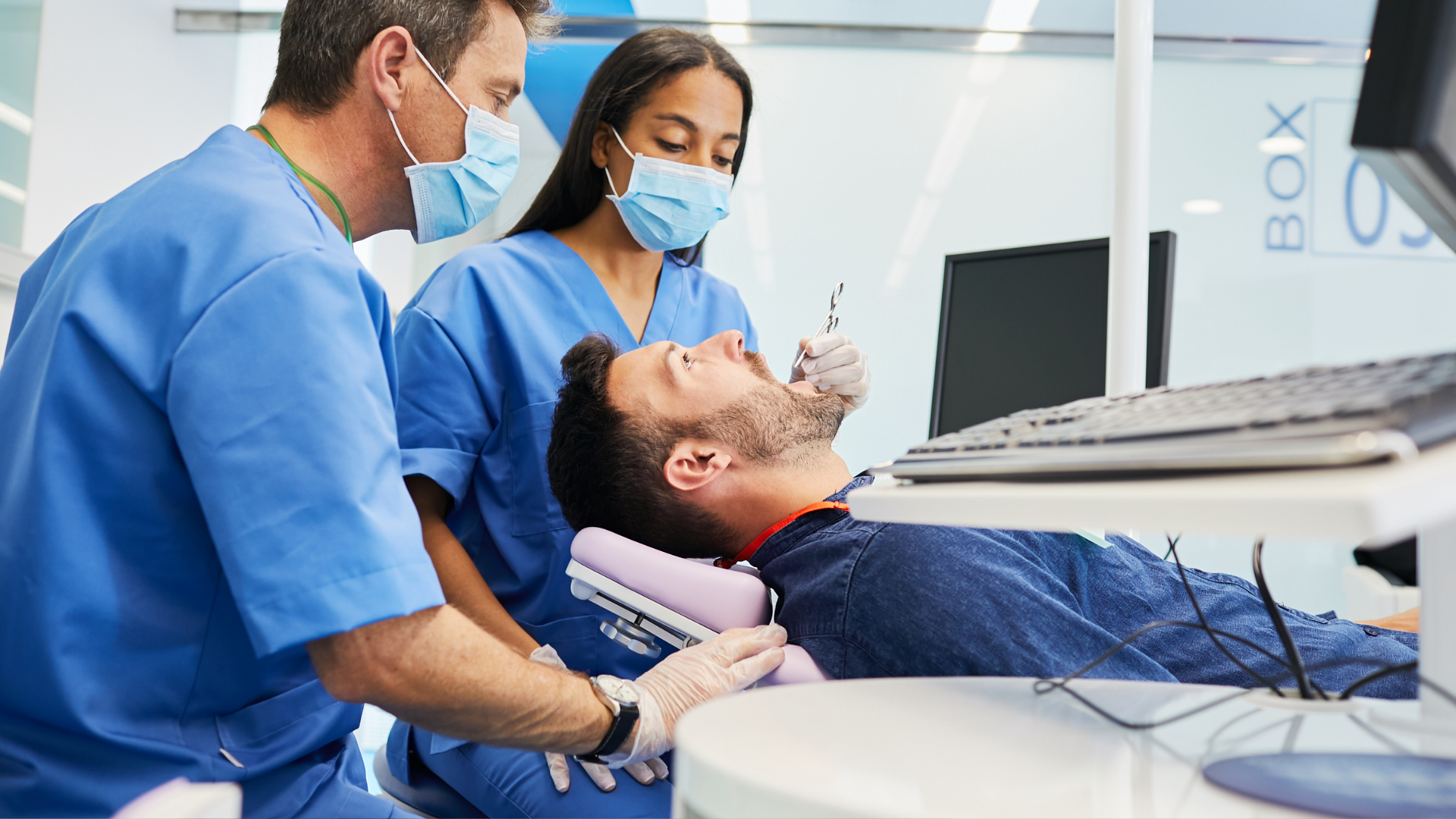 Dental team examining a patient reclined in a chair, with a laptop and monitor nearby.