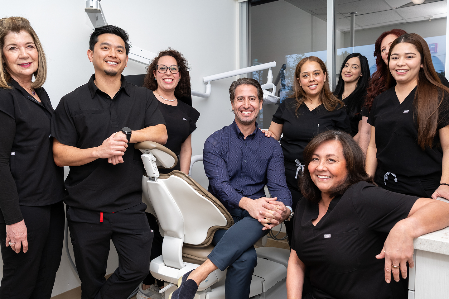 Group of smiling dental staff in black uniforms gathered around a reclining dental chair in a clinic