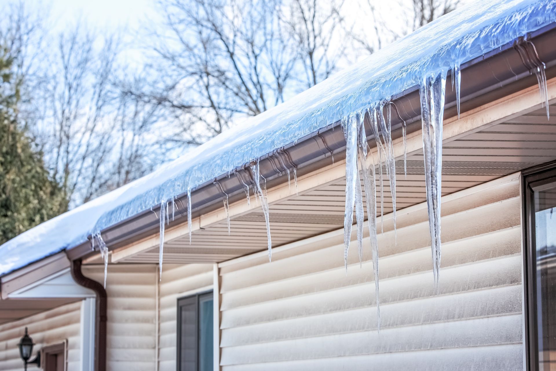 Icicles hanging from a roof's edge of a beige house on a winter day. Blue sky, bare trees.