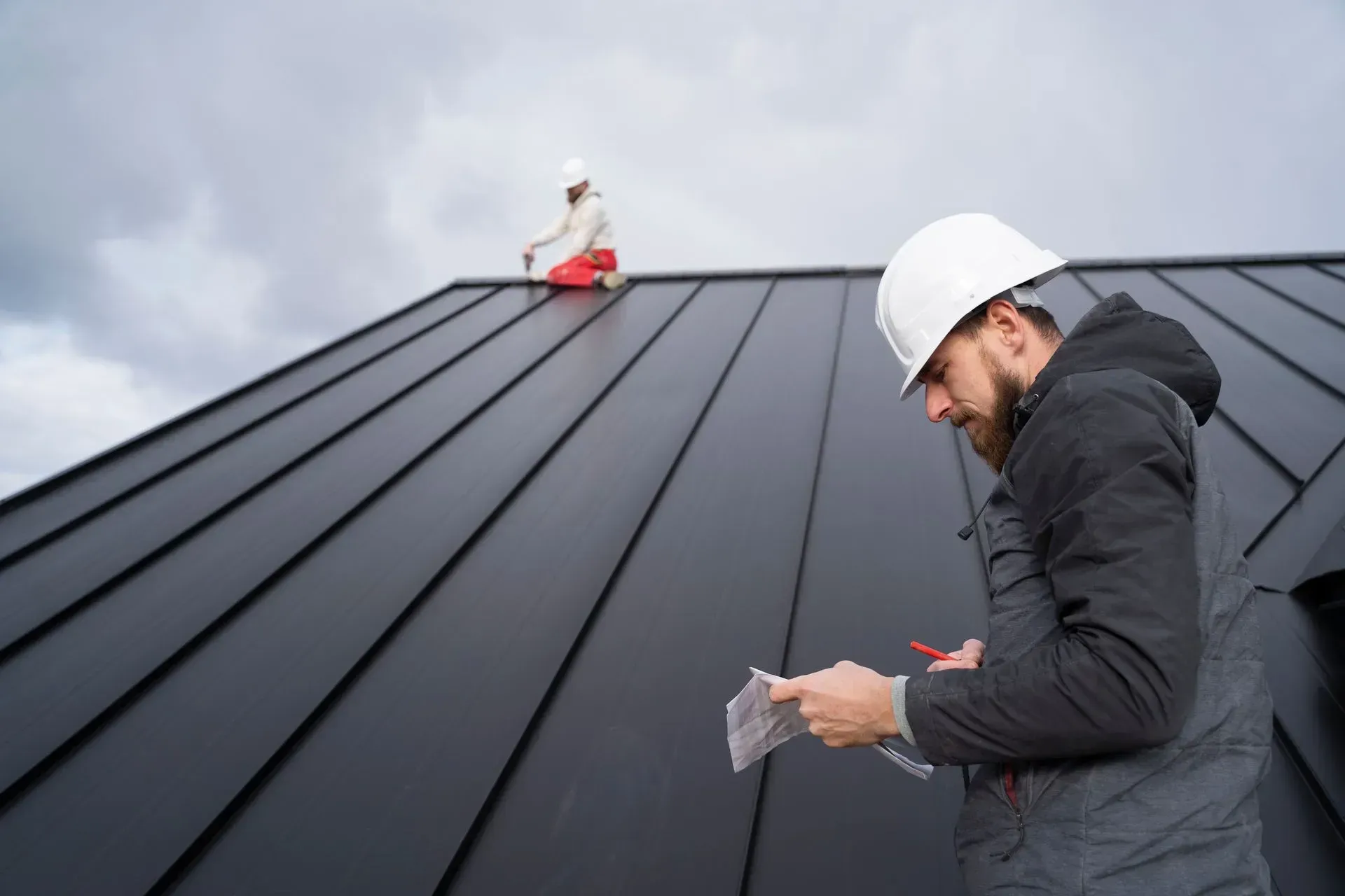 Two roofers on a black metal roof; one working, the other examining paperwork. Overcast sky.