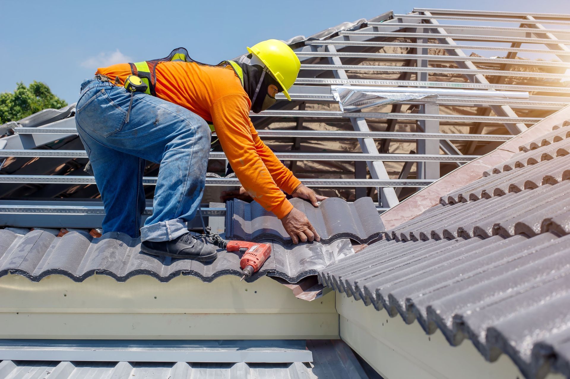 Roofer in orange shirt and yellow hard hat installing tiles on a roof, sunny day.