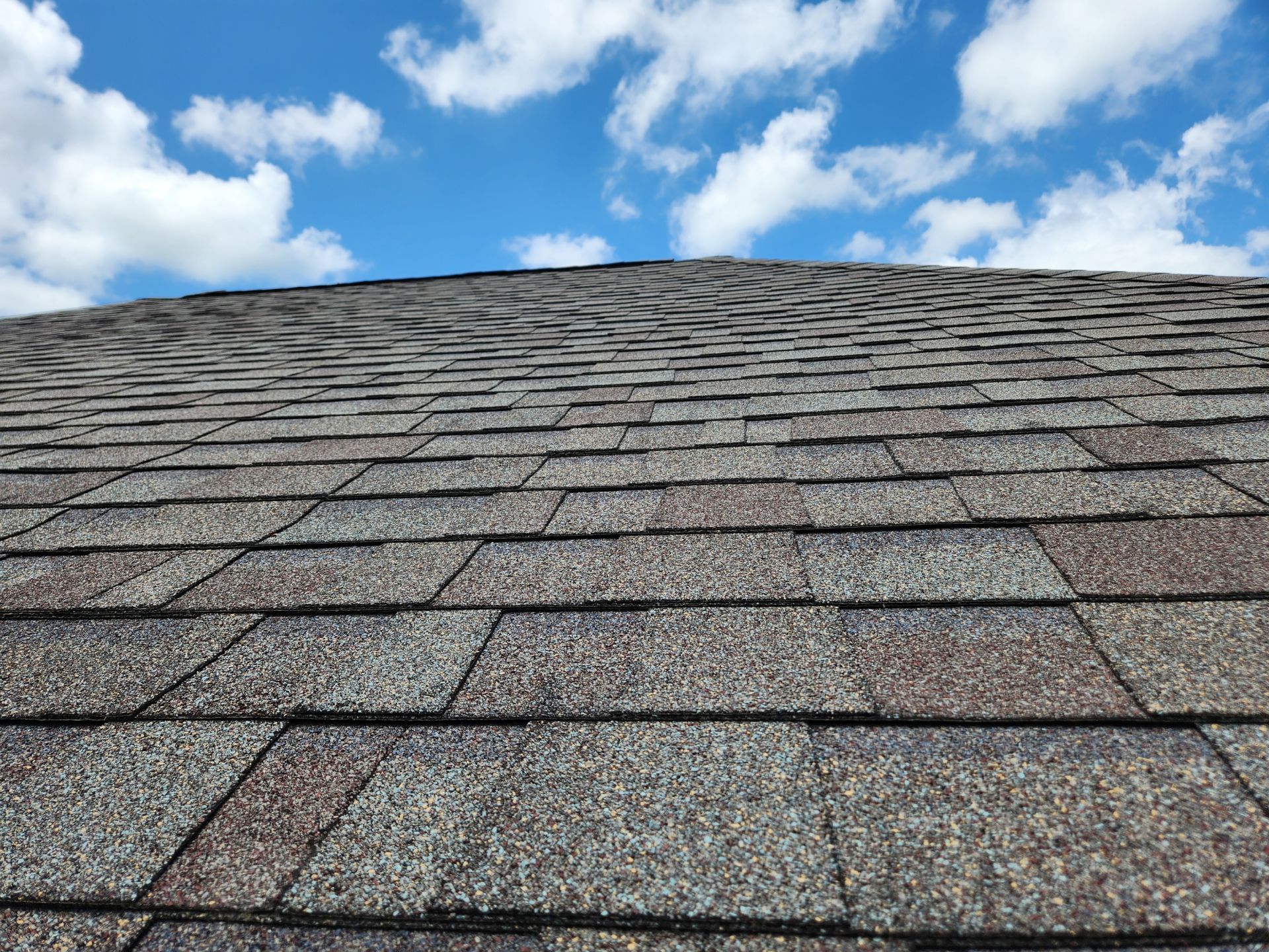 Roof covered in gray and brown shingles against a blue sky with white clouds.