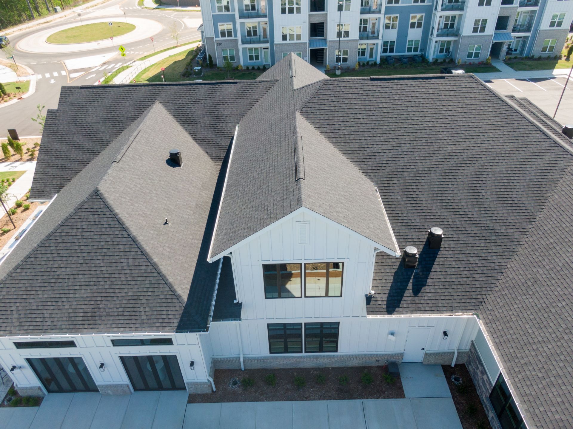 Aerial view of a white building with a black shingled roof, set near a roundabout and apartments.