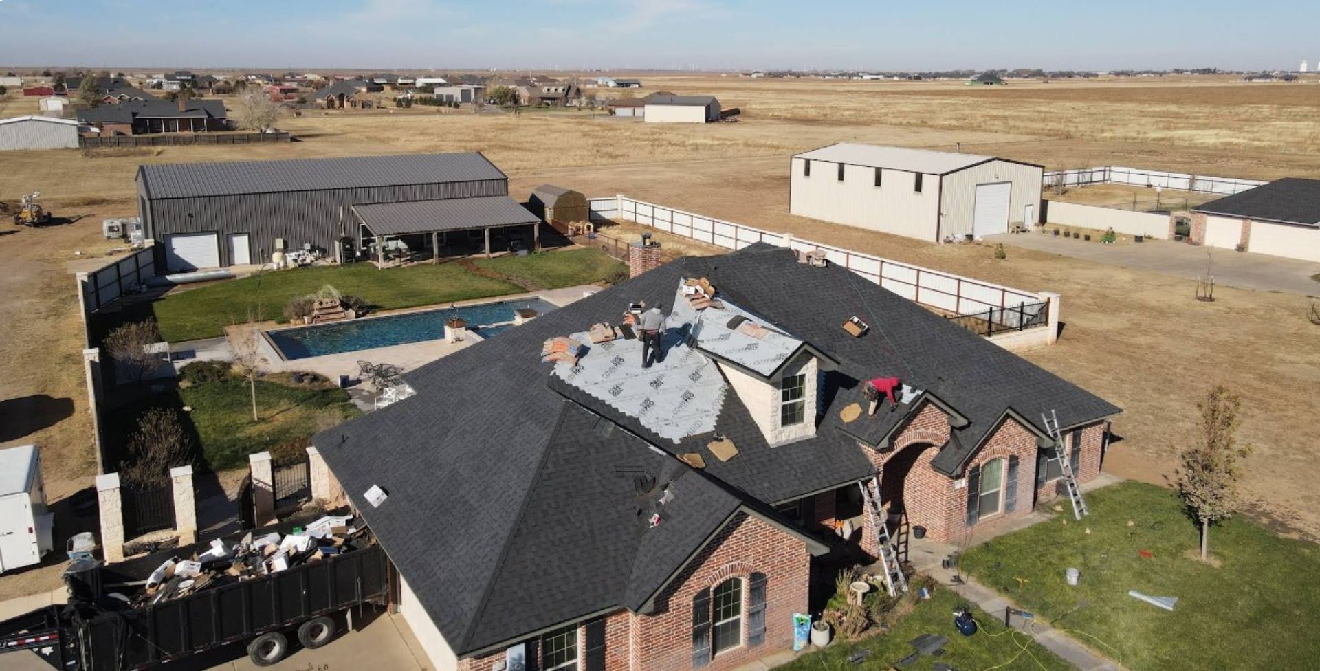 Aerial view of a home with roof damage, roofing crew, debris, pool, and surrounding rural landscape on a sunny day.