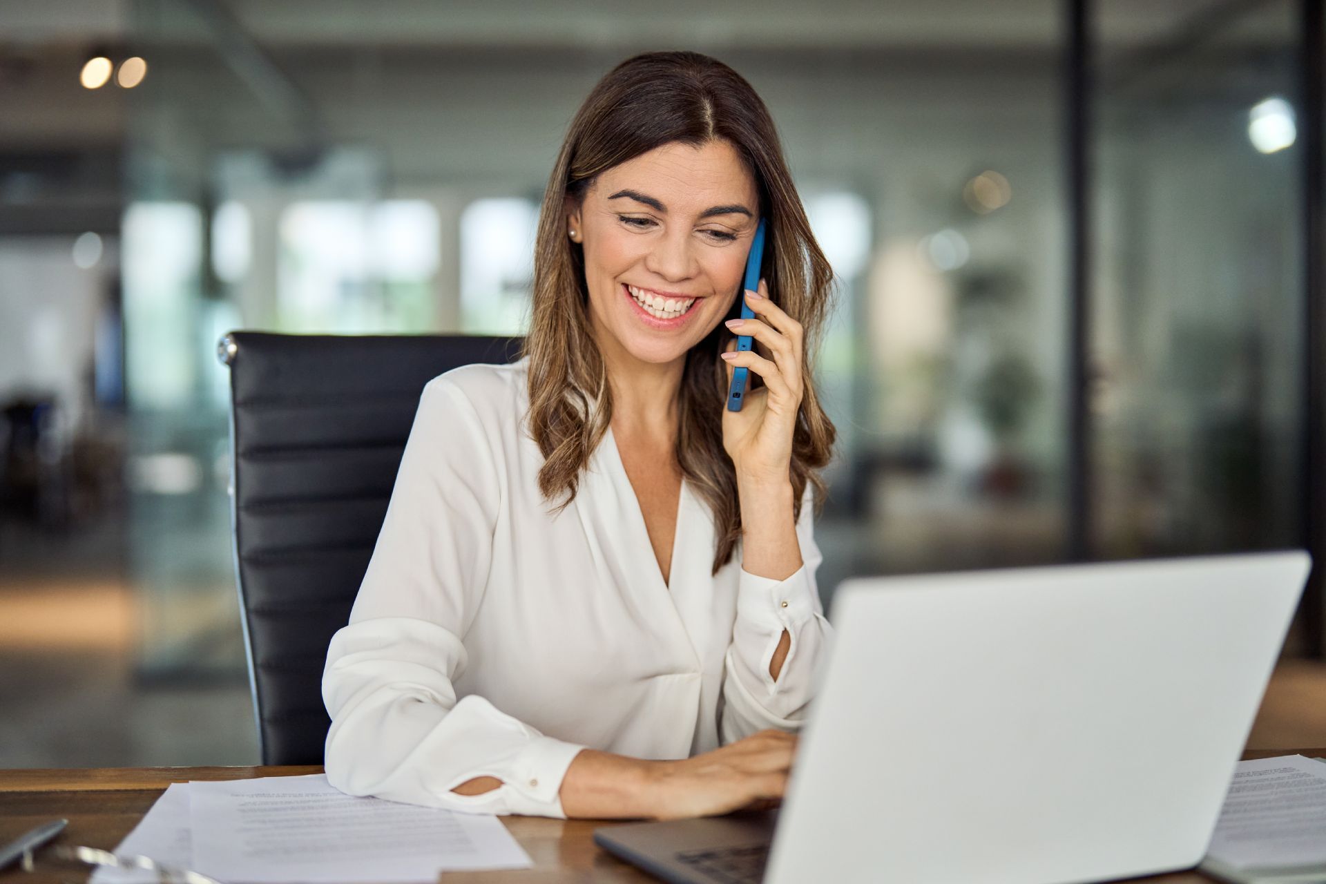 Woman smiling, on phone, working at desk with laptop in modern office setting.