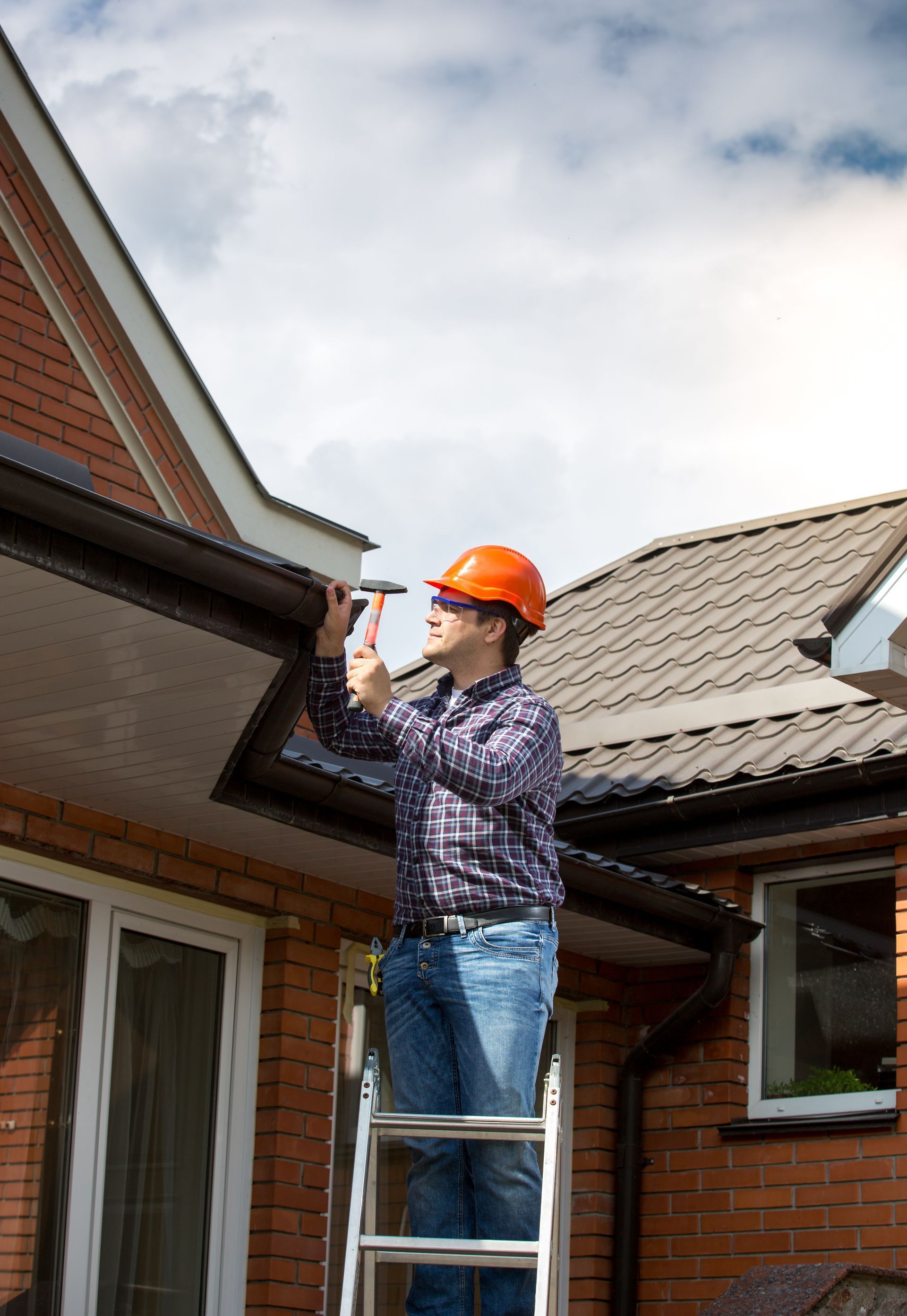 Person in hard hat on a ladder, installing a gutter on a brick house.