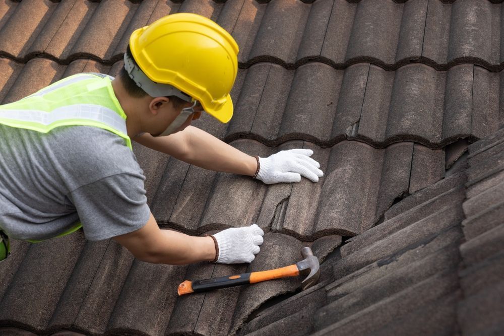 Roofer in yellow hard hat fixes tile roof with hammer, wearing gloves and safety vest.