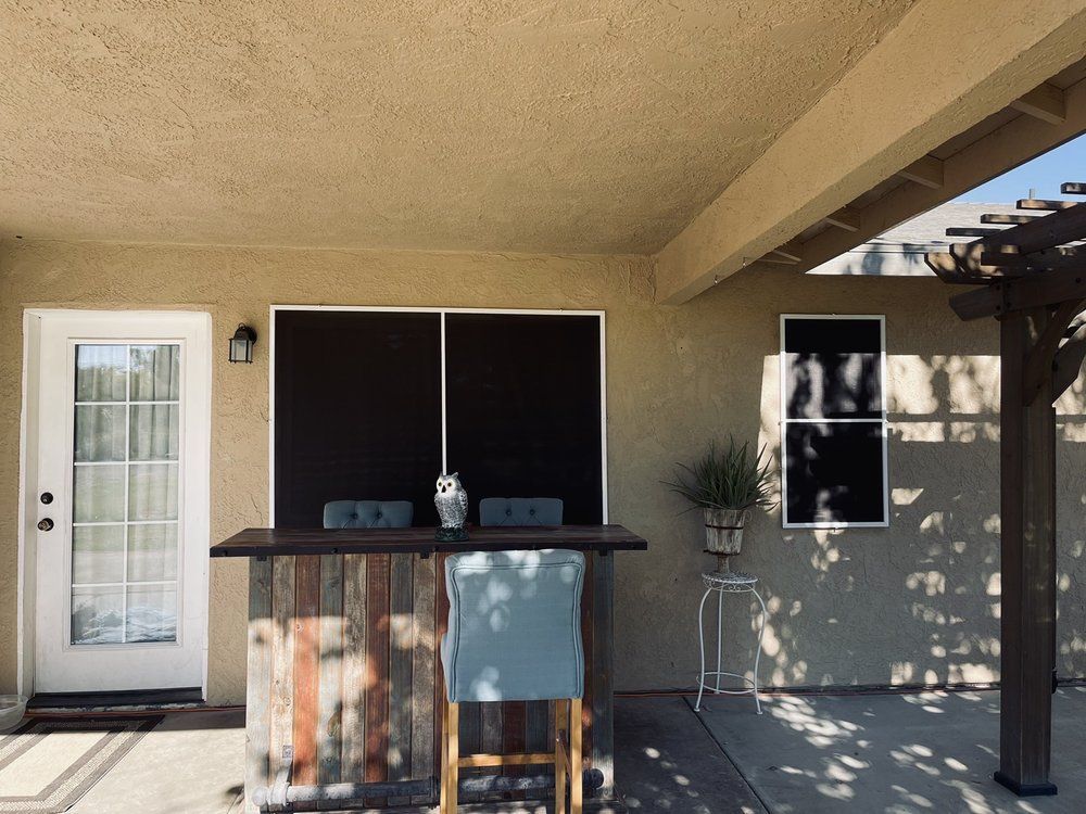 Outdoor bar with a rustic wooden facade, black window coverings, and a white door on a patio.