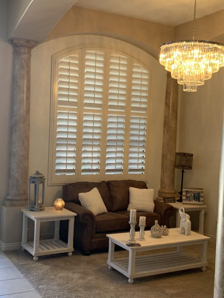 Cozy living room with brown sofa, white shutters, chandelier, and side tables; neutral color scheme.