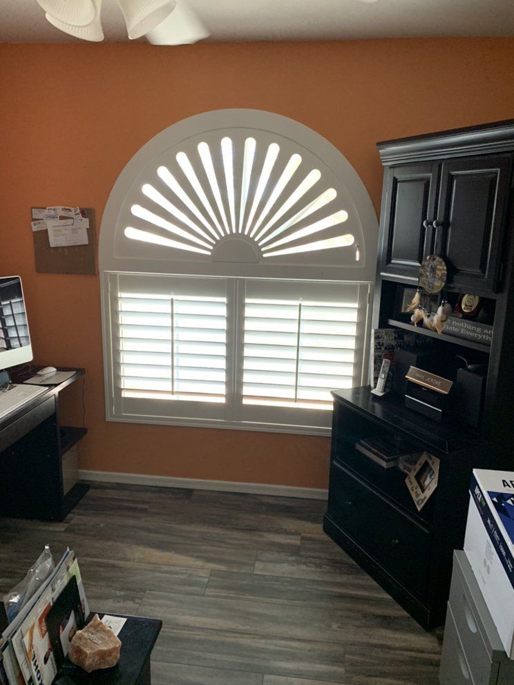 White shutters with arched sunburst over a window in an orange-walled room. Black cabinet to the right.