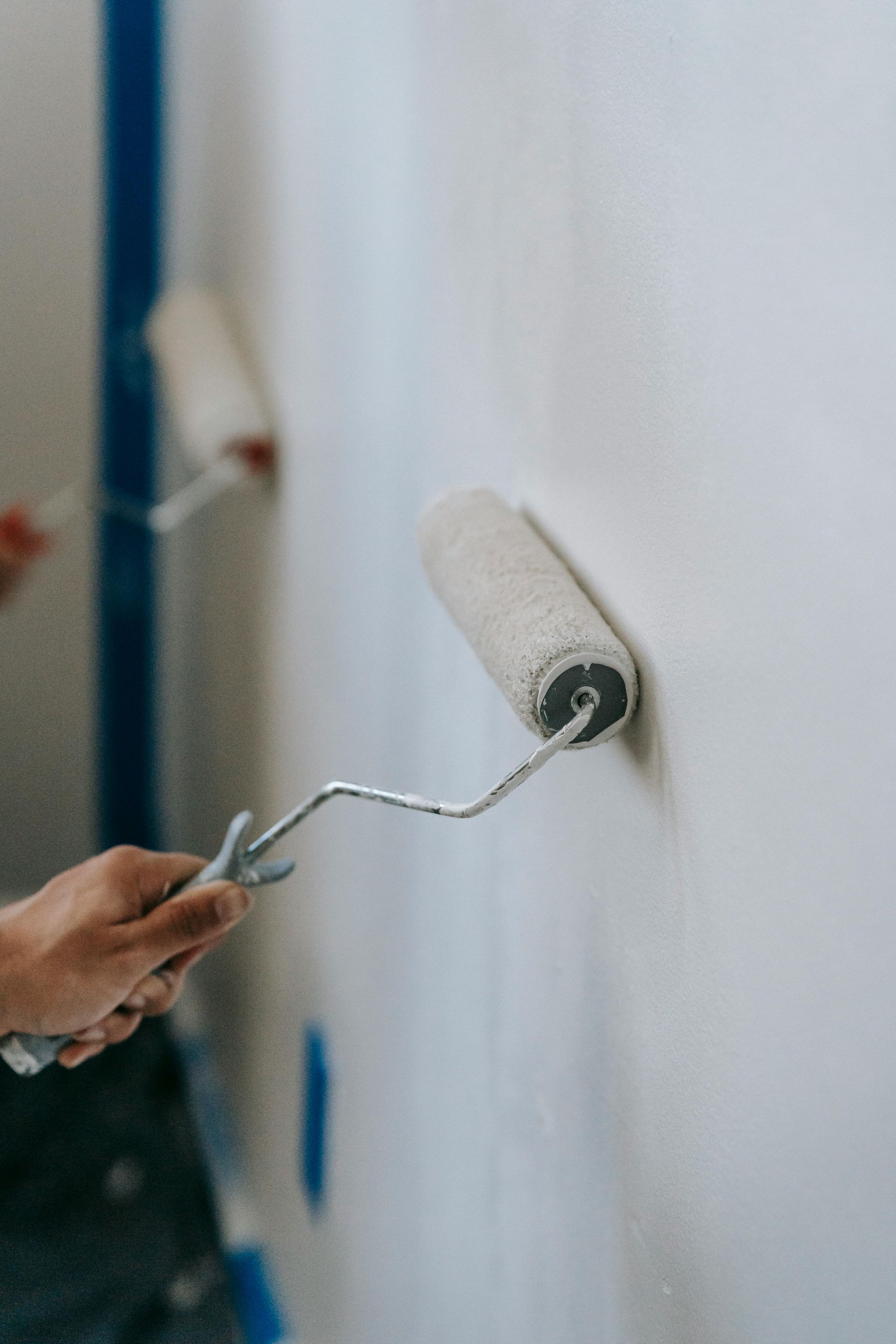 Hand rolling white paint onto a wall; blue tape and another roller in the background.