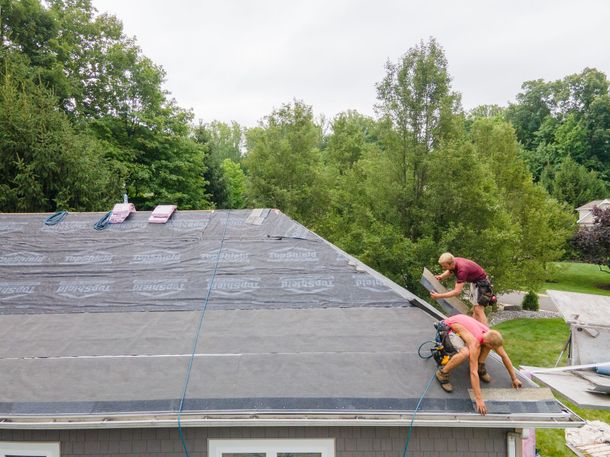 Roofers working on a house roof, attaching roofing material, surrounded by trees.