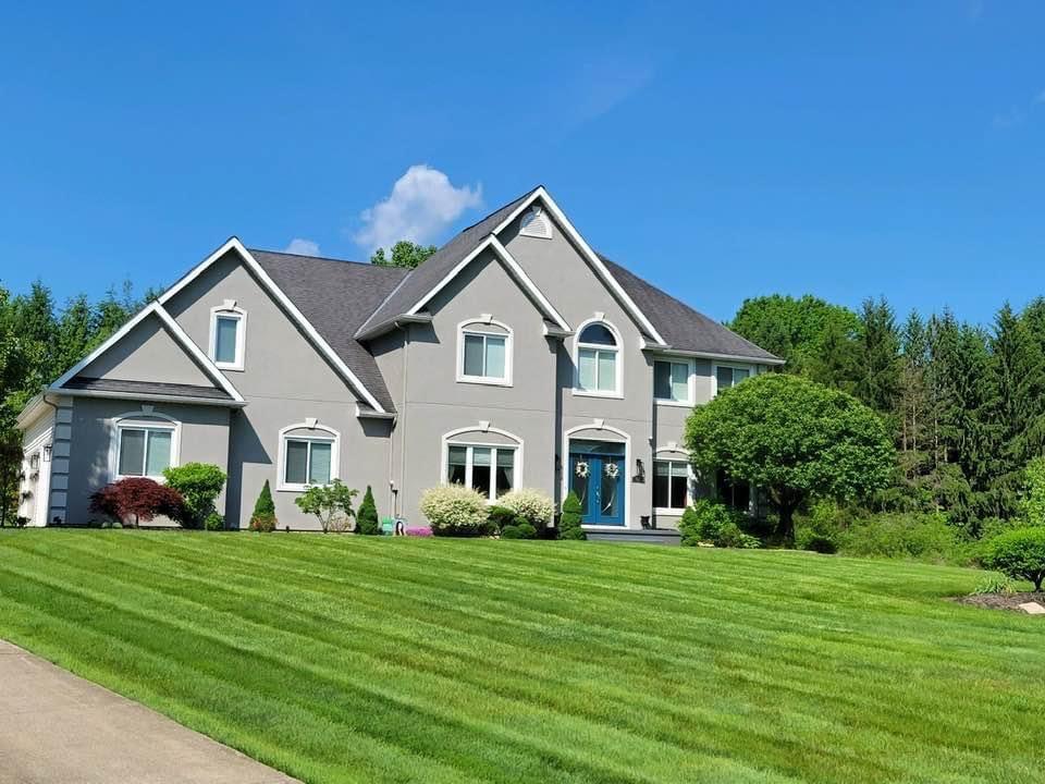 Gray two-story house with green lawn under a blue sky; trees in the background.