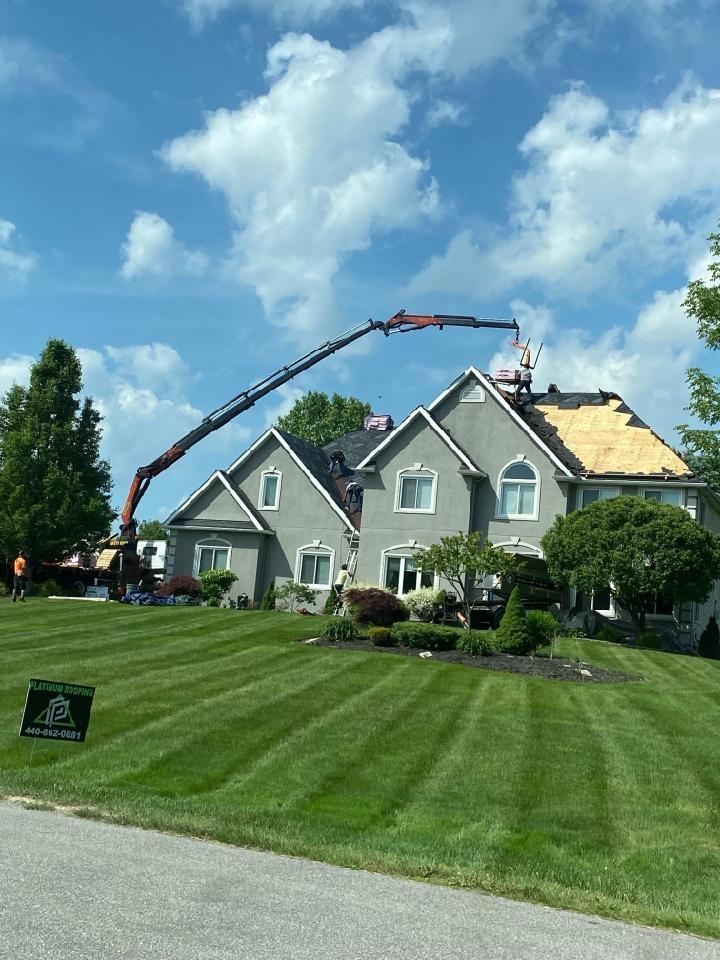 Crane reaching a house roof, roofers working. Green lawn, blue sky with clouds.
