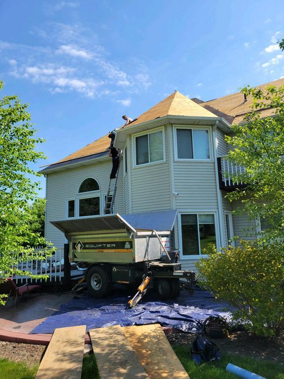 Man on ladder near house; blower machine at base, blue tarp, sunny day.