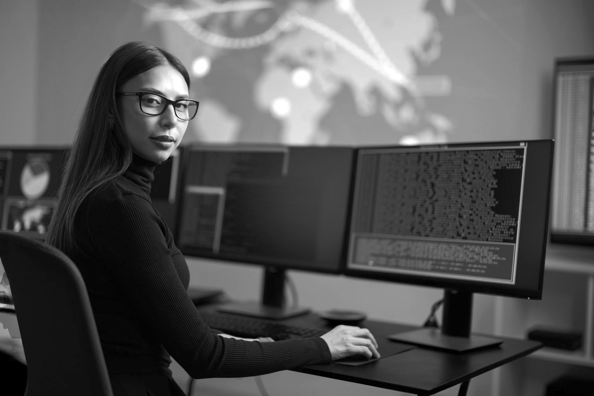 Woman at computer monitors, analyzing data. World map graphic in background. Black and white.