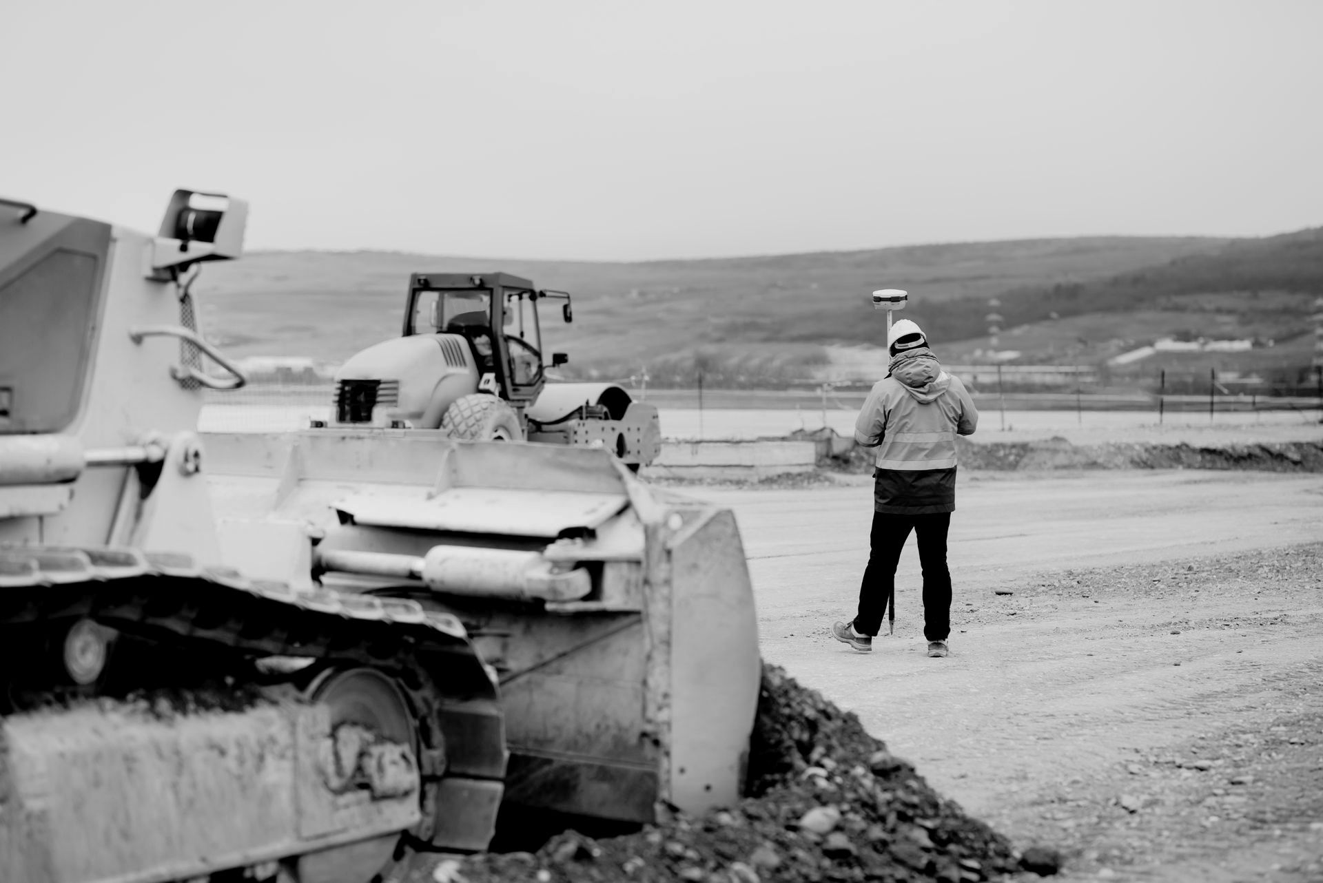A construction worker uses surveying equipment on a job site with heavy machinery, a cloudy day, and a distant landscape.