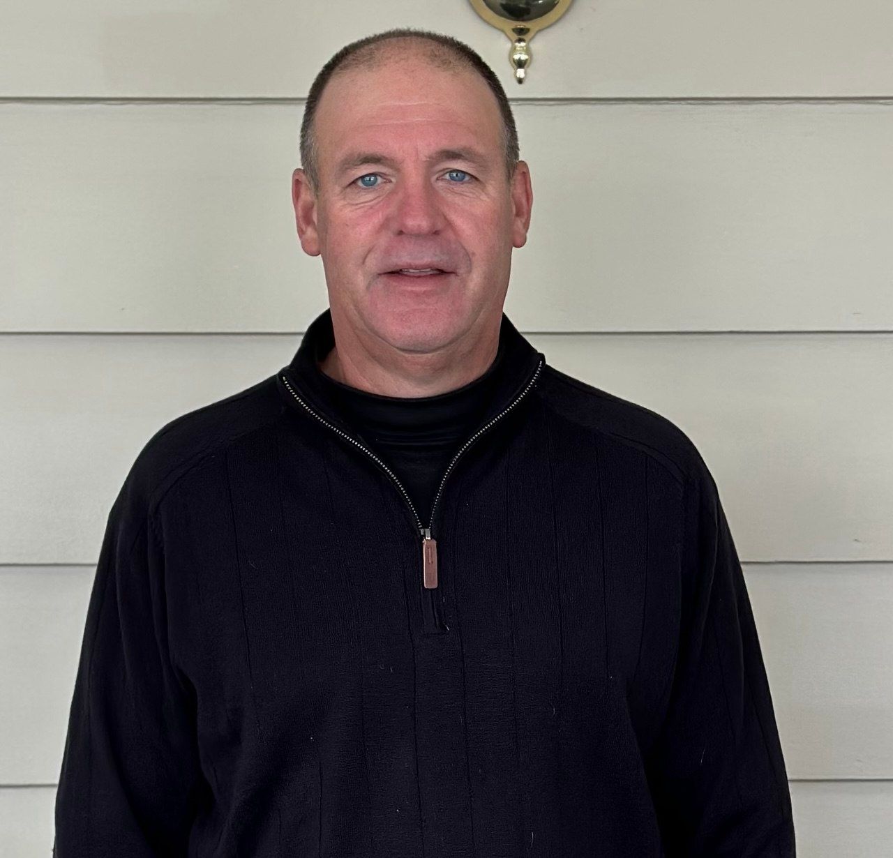 A man in a black sweater stands in front of a white wall