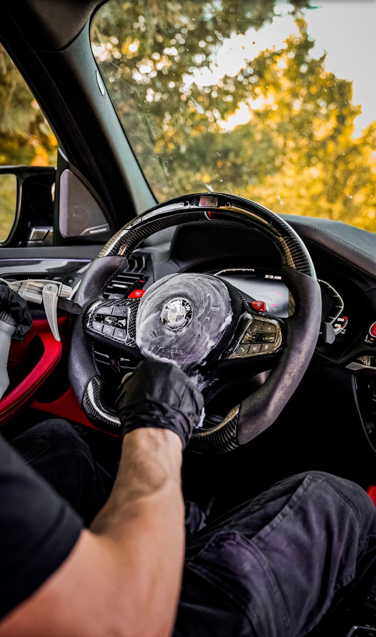 Person in a car holding a steering wheel with black gloves. Background is trees, interior red.