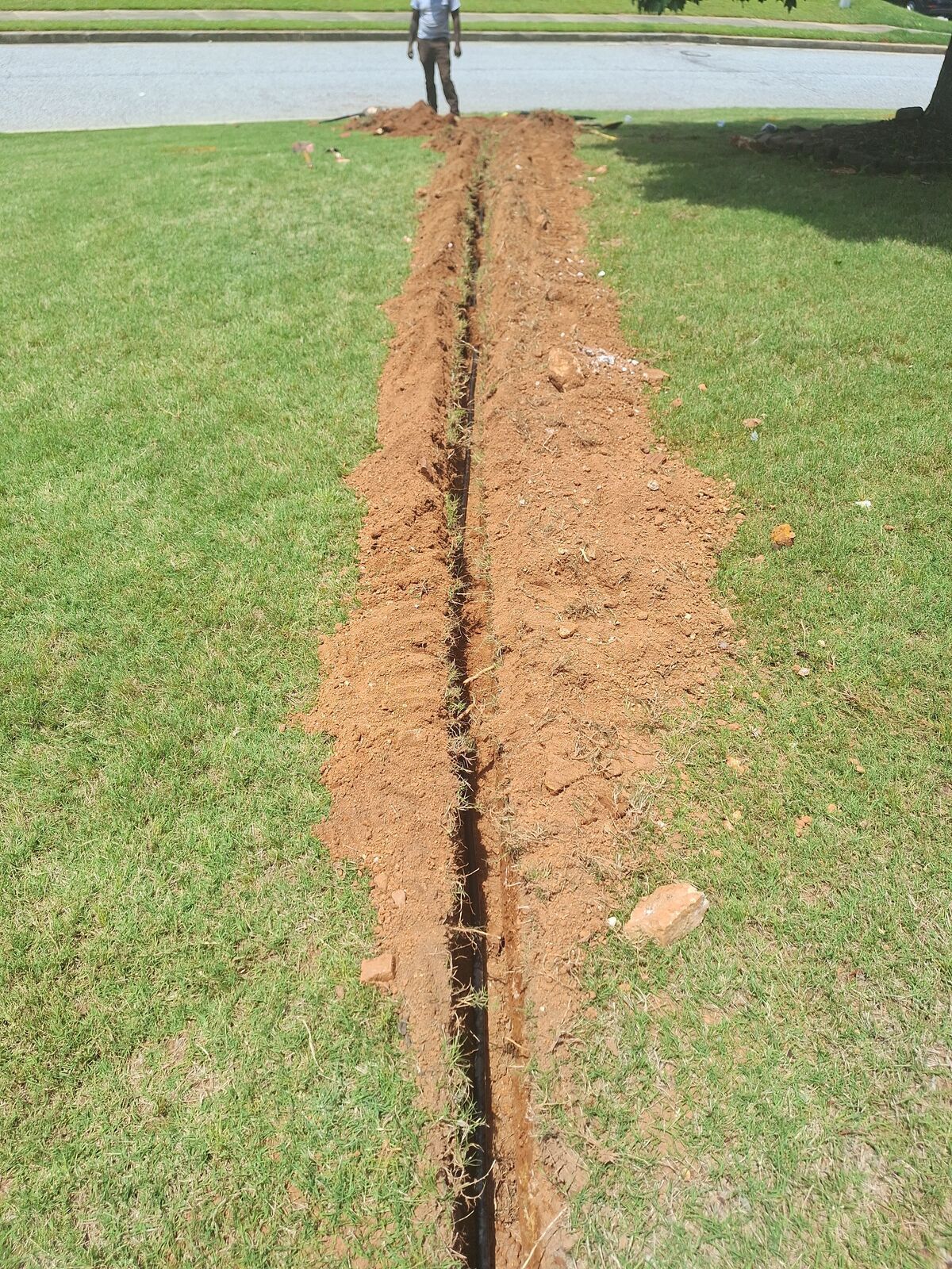 Long narrow trench dug in green grass with a person standing at the end near a road.
