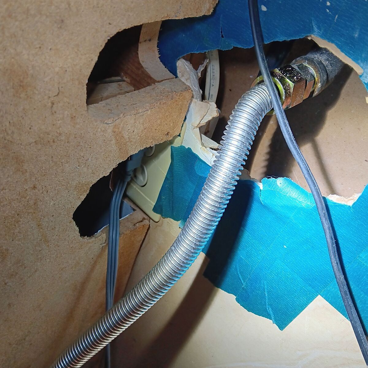 Close-up of a damaged wall with a metallic, flexible tube and electrical wiring. Blue tape secures the edges.