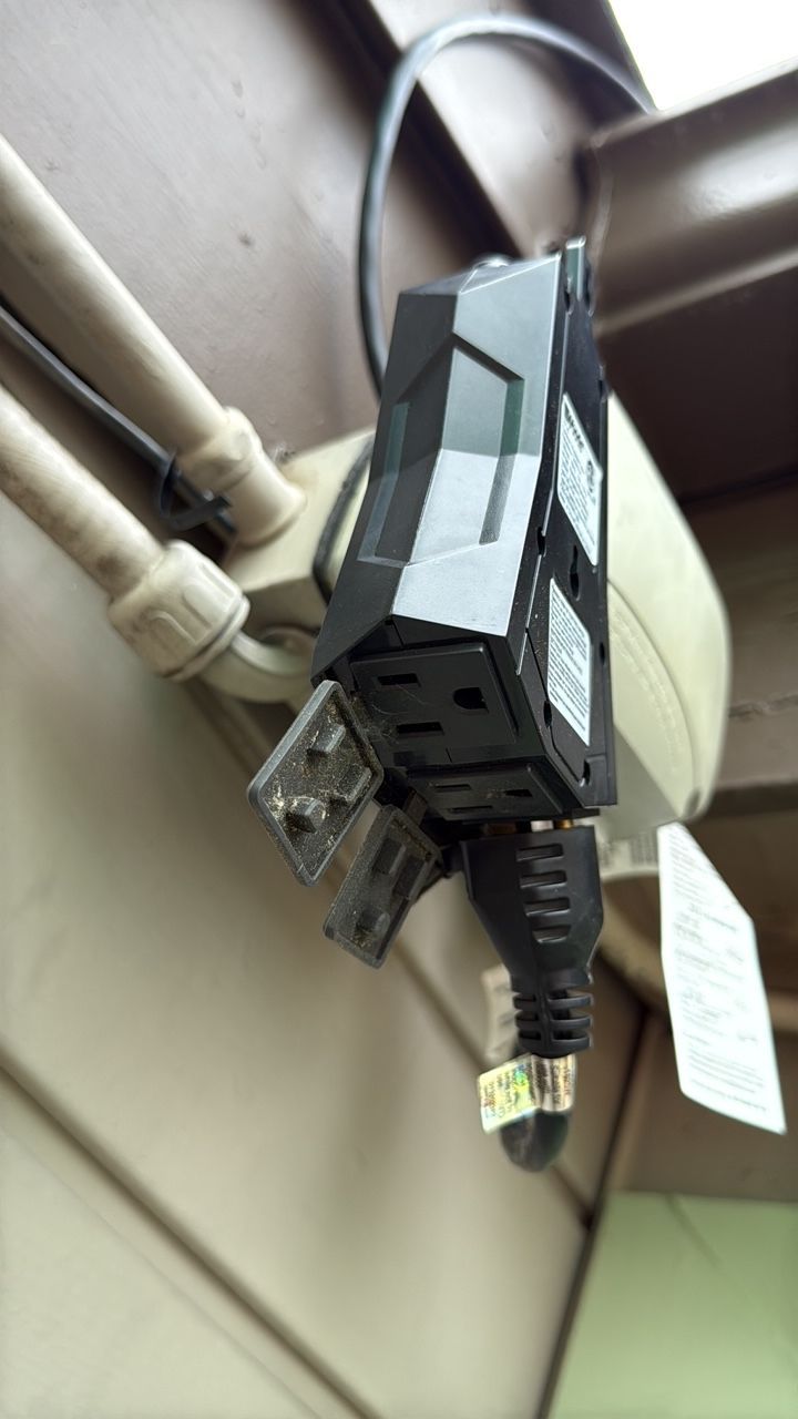 Black power strip with three outlets plugged into a tan wall outlet, mounted on a building's exterior.