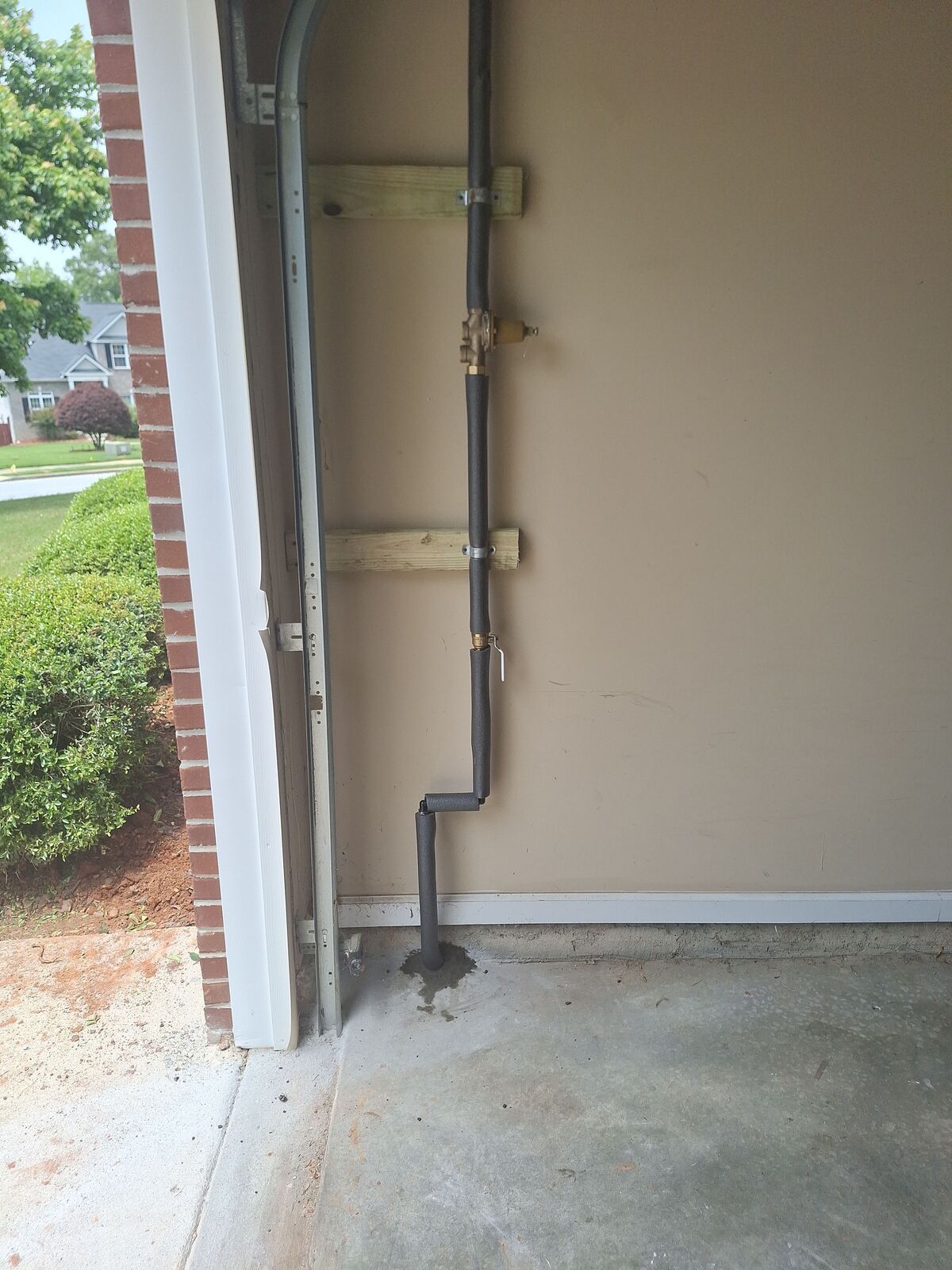 Black pipe running vertically along beige garage wall, secured with wooden supports. Concrete floor.