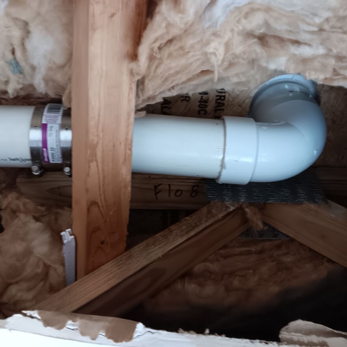 White PVC pipe and elbow in an attic, connected with a metal clamp, surrounded by insulation.