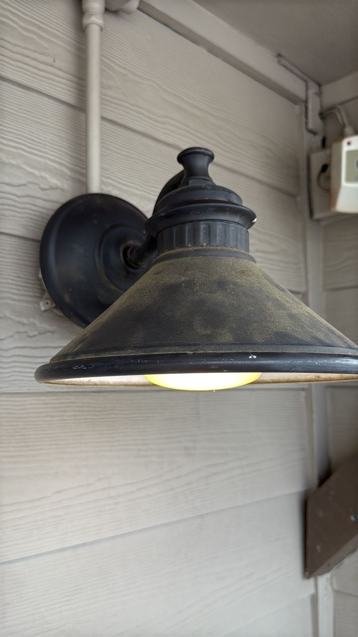 Black outdoor light fixture on a beige siding wall; light is on.