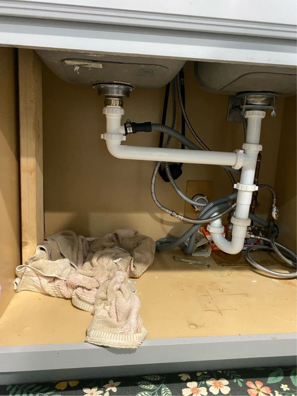 Underside of a double sink cabinet with white plumbing pipes, wires, and a cloth.