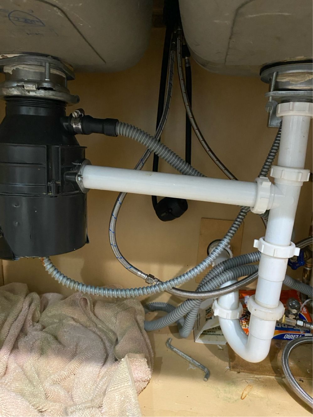 Under-sink plumbing with a garbage disposal and drain pipes, against a light brown cabinet.