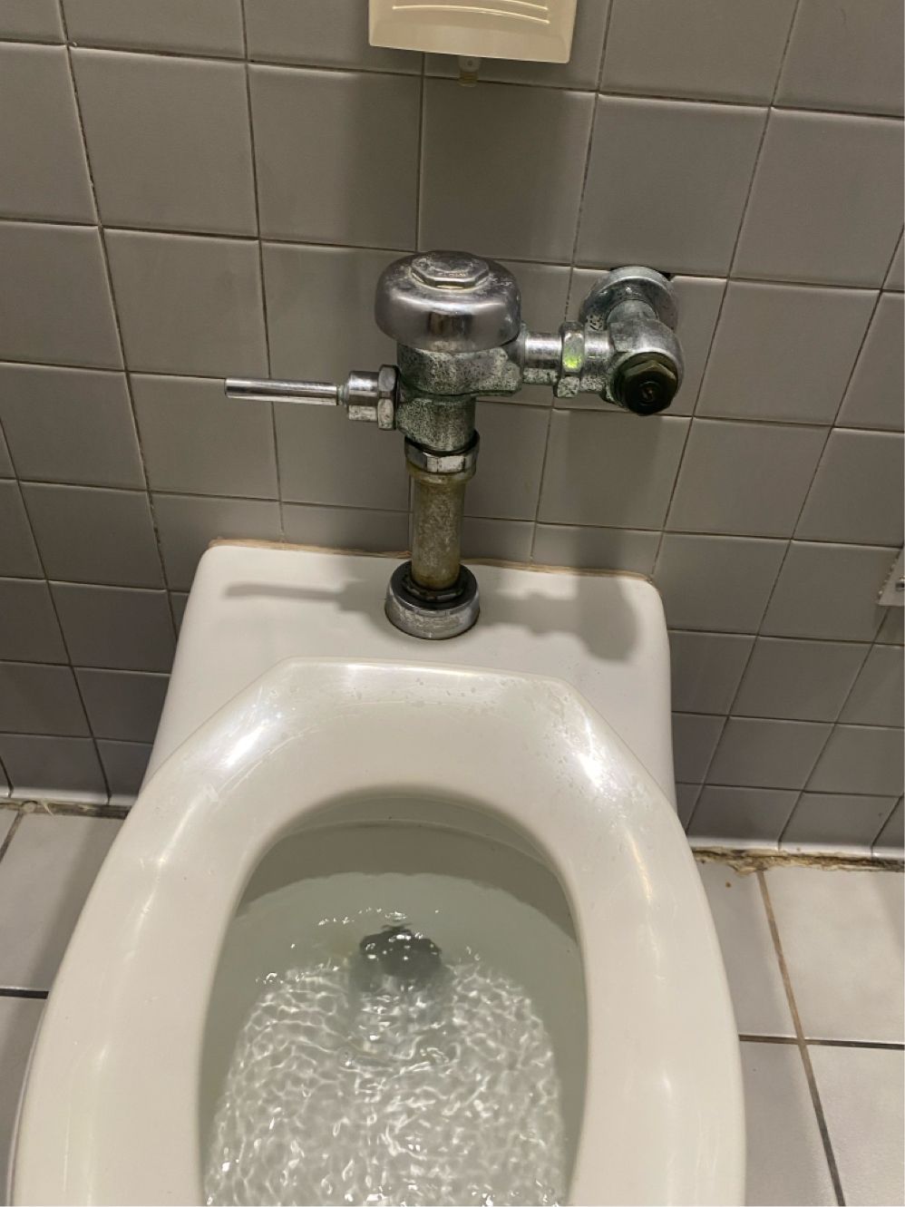 Toilet with silver flush valve and running water in a tiled restroom.