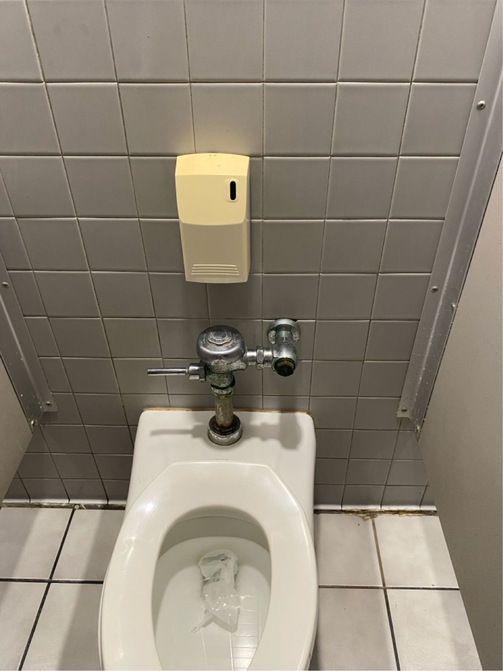 Toilet in a public restroom with a wall-mounted soap dispenser and tiled wall.