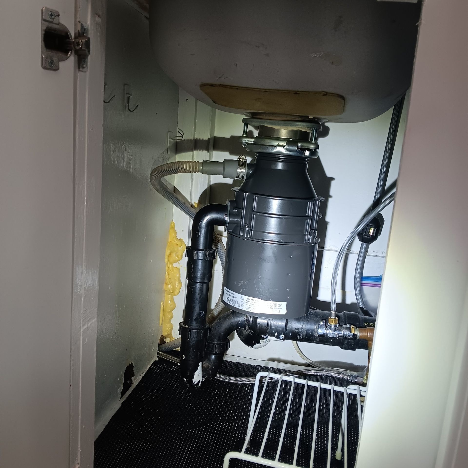 Under-sink view of a garbage disposal unit with connected pipes, inside a white cabinet.