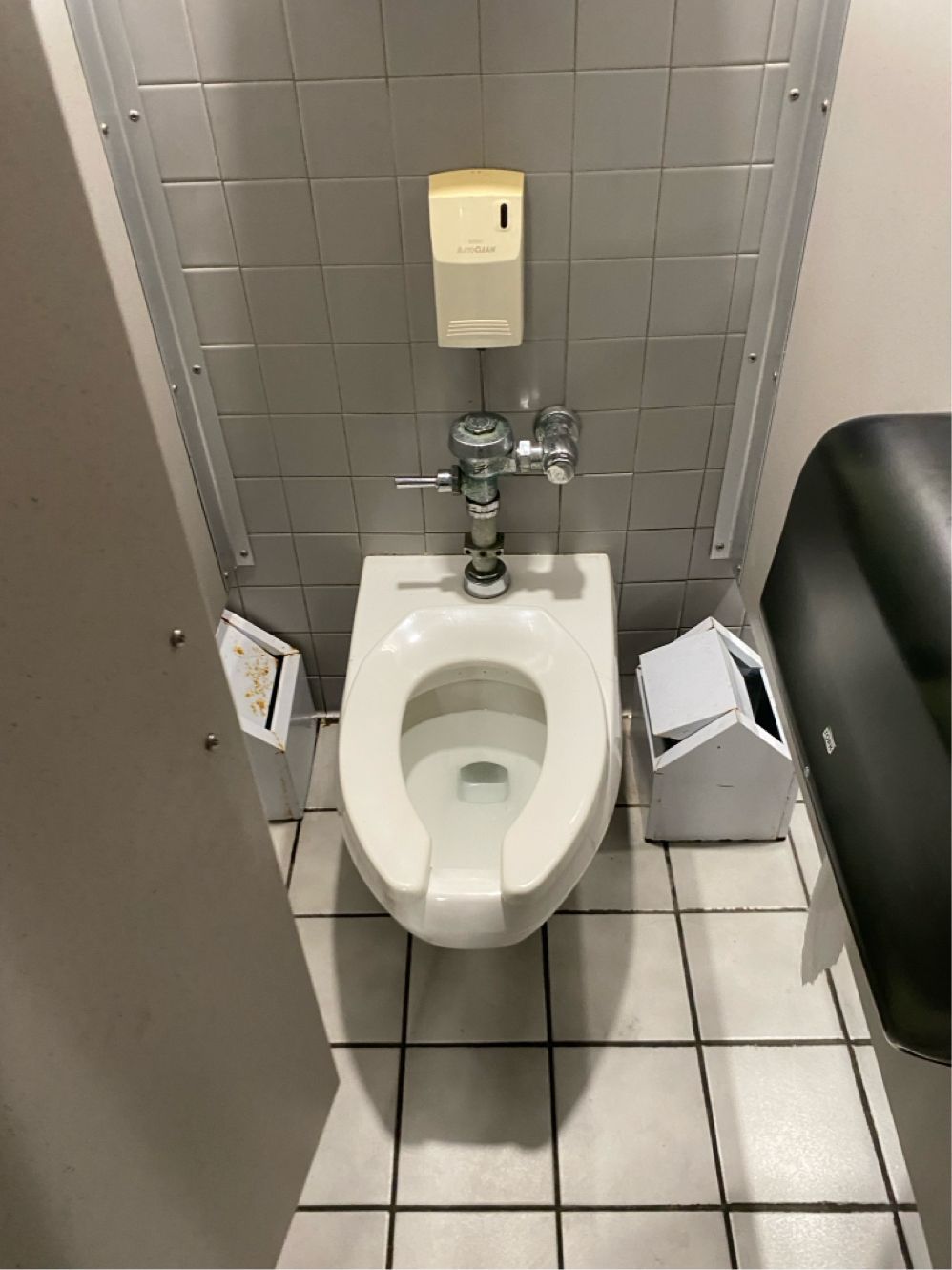 Toilet in a public restroom stall with a paper towel dispenser, and trash receptacles.