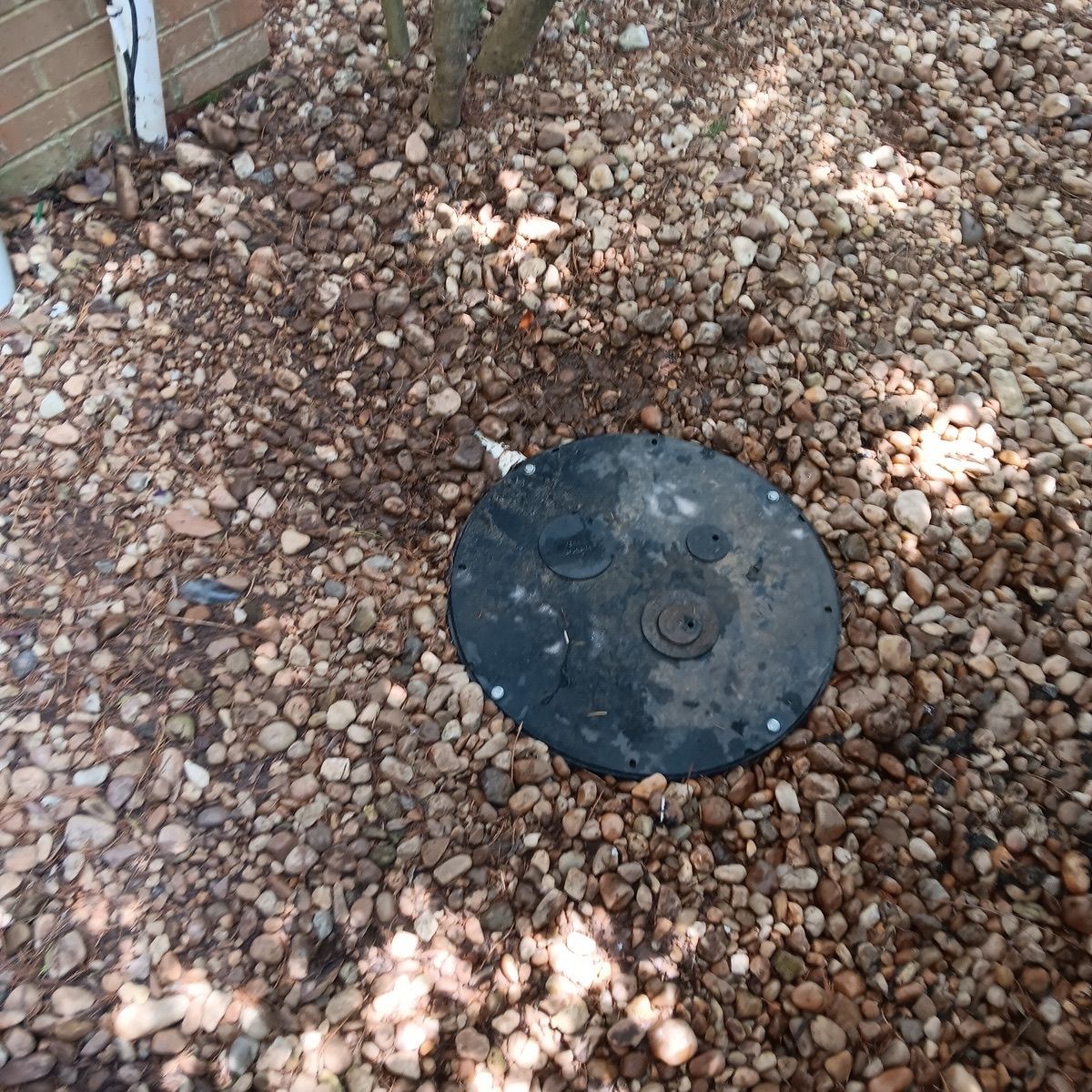 Black circular access cover in gravel next to a brick wall.