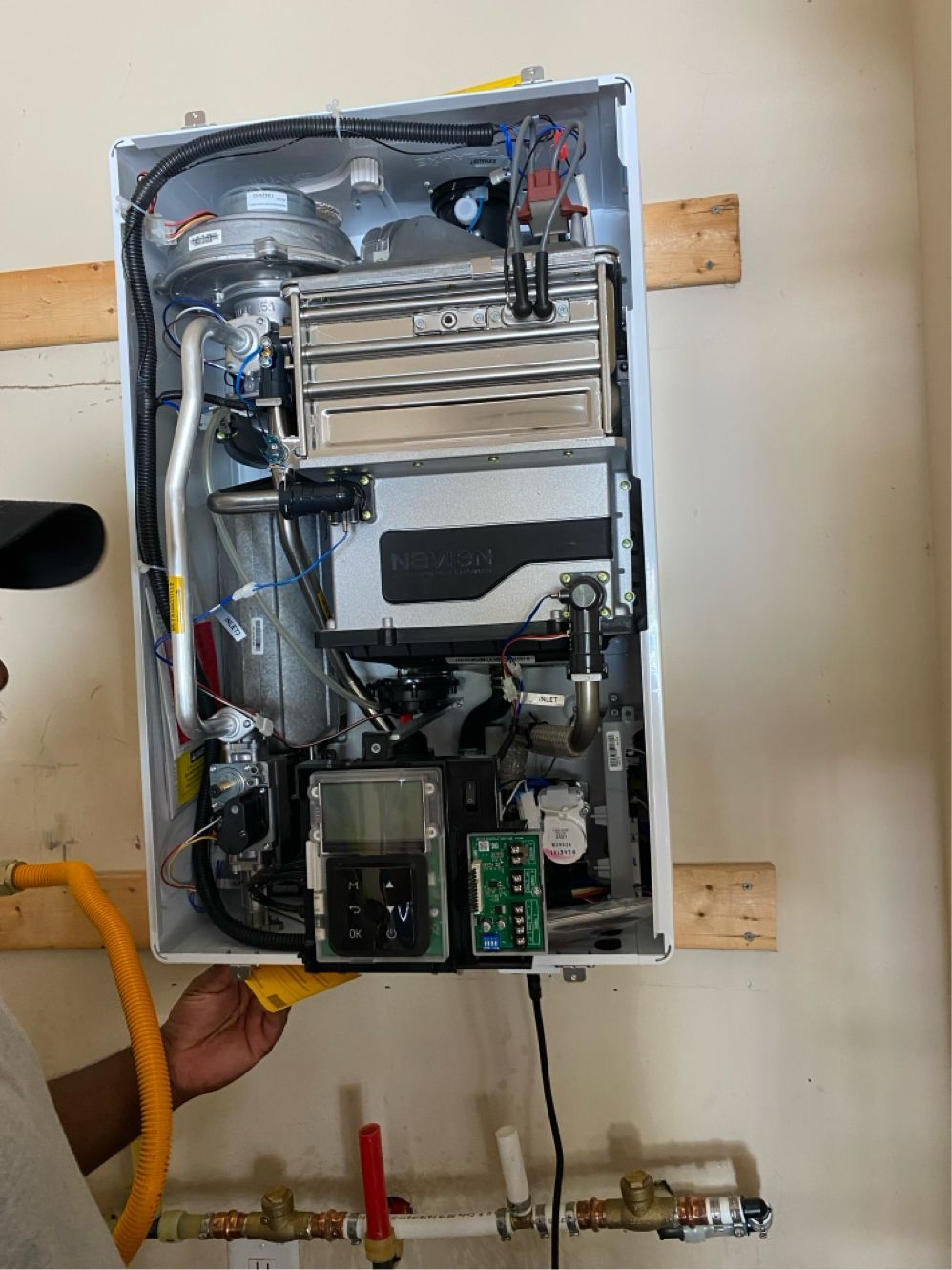 A gas water heater with its components exposed is held by a person. Mounted on a wall, with water pipes visible.