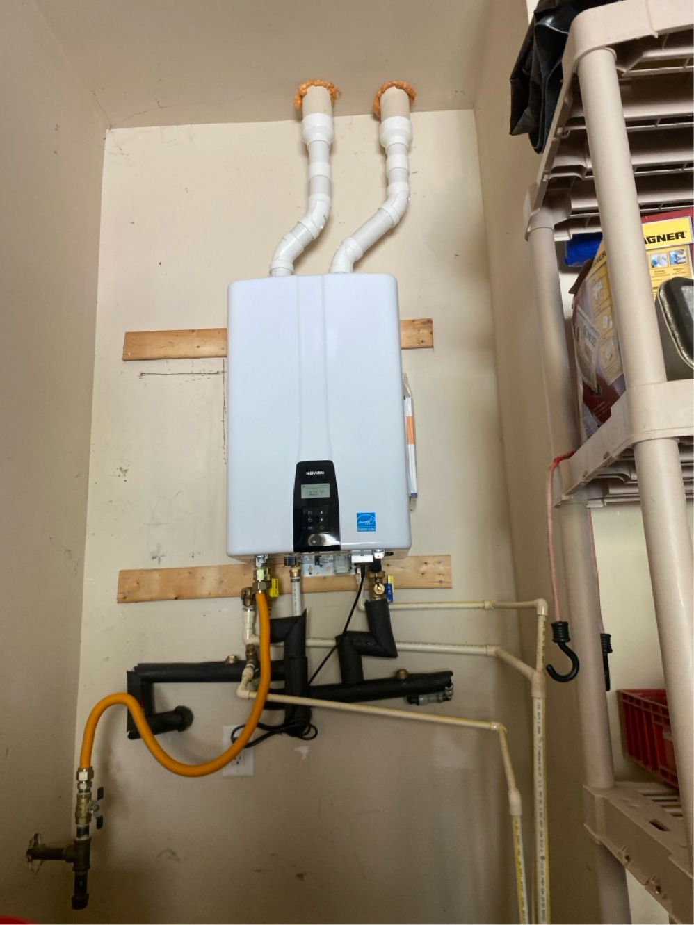 White tankless water heater mounted on a beige wall with venting, pipes, and gas line. Shelving is to the right.