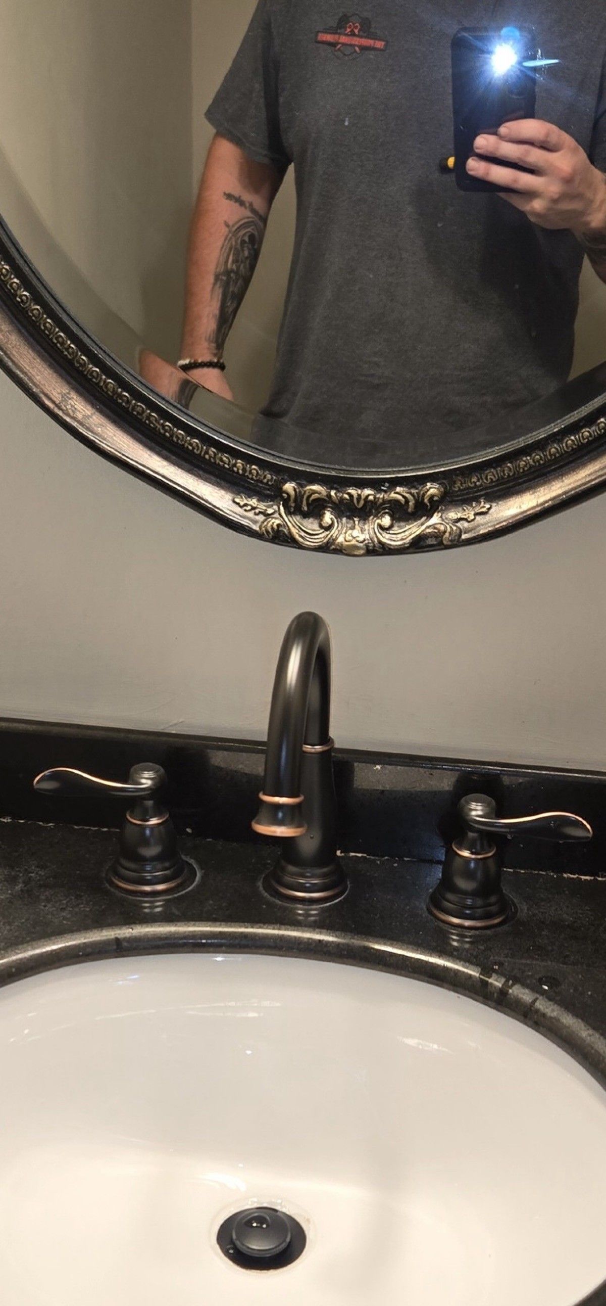Person taking photo of sink in mirror. Bronze faucet and handles. Dark counter and white basin.