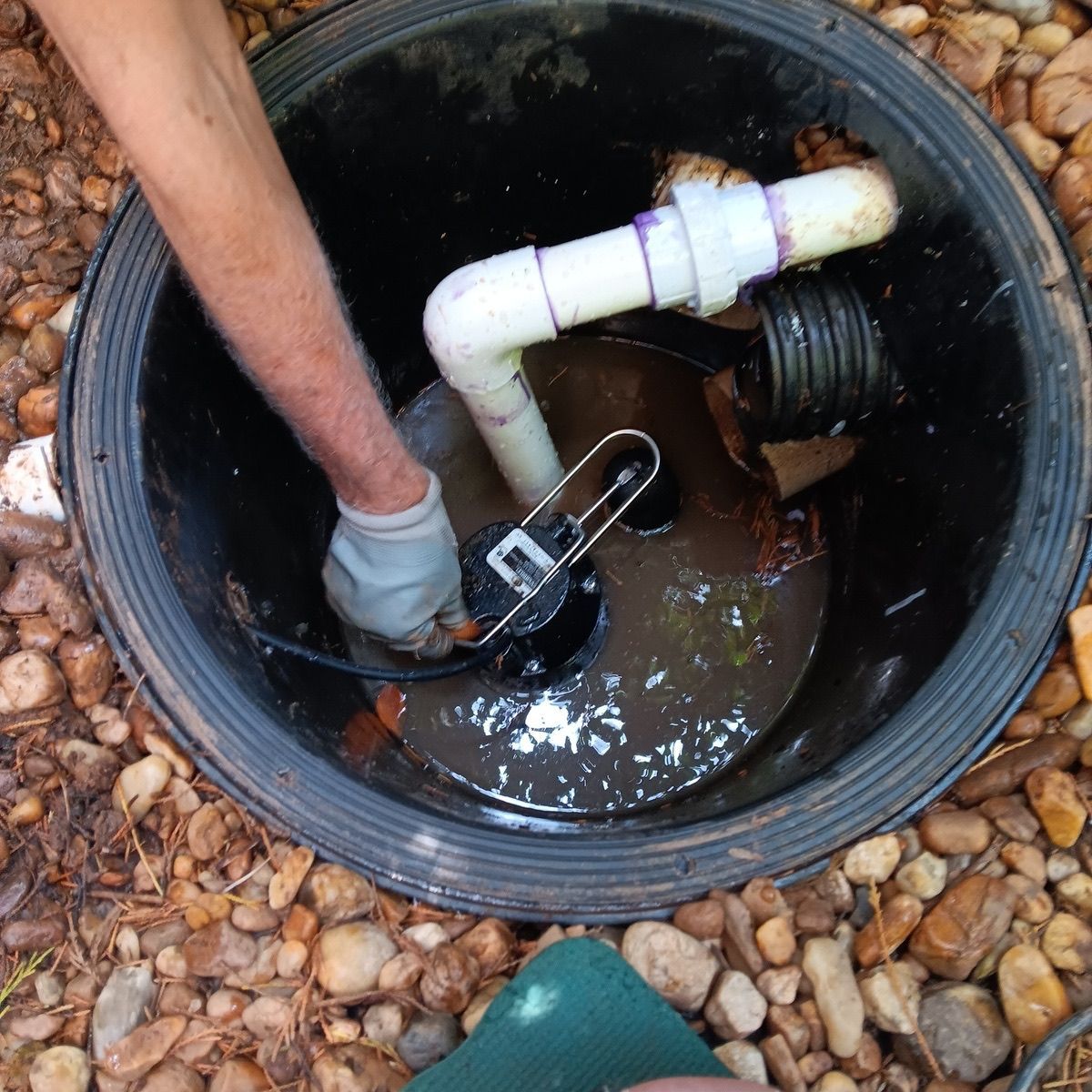 Person with glove working on a sump pump in a black basin filled with water, surrounded by rocks.