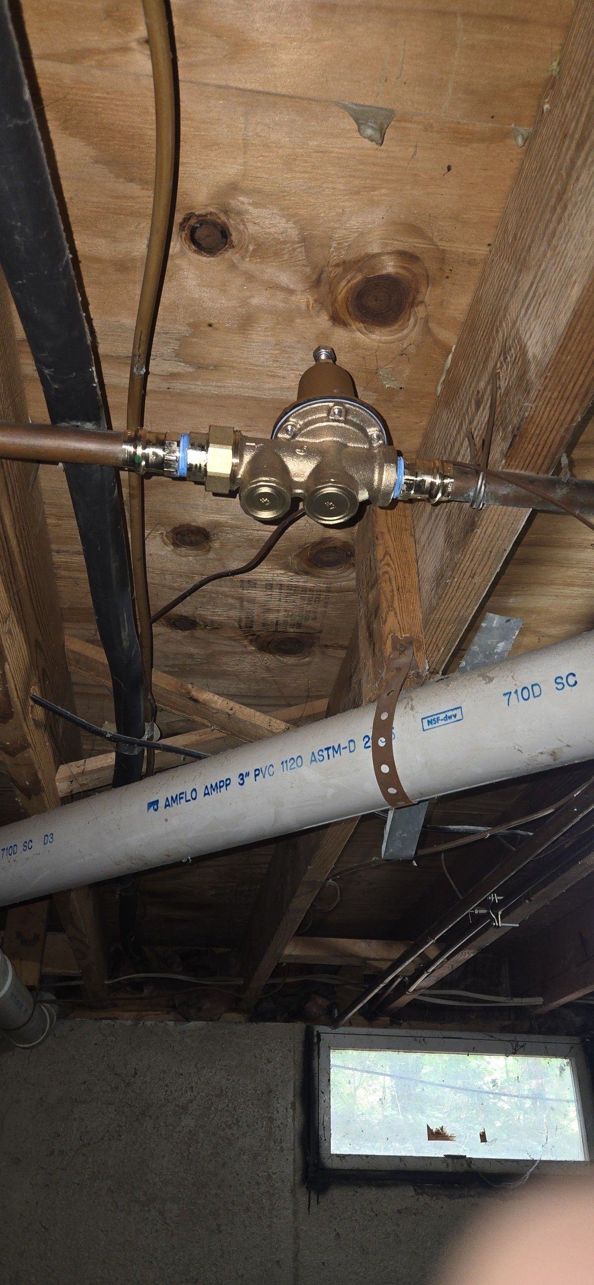 Water pressure regulator on copper pipes in a basement. White PVC pipe and wooden ceiling visible.