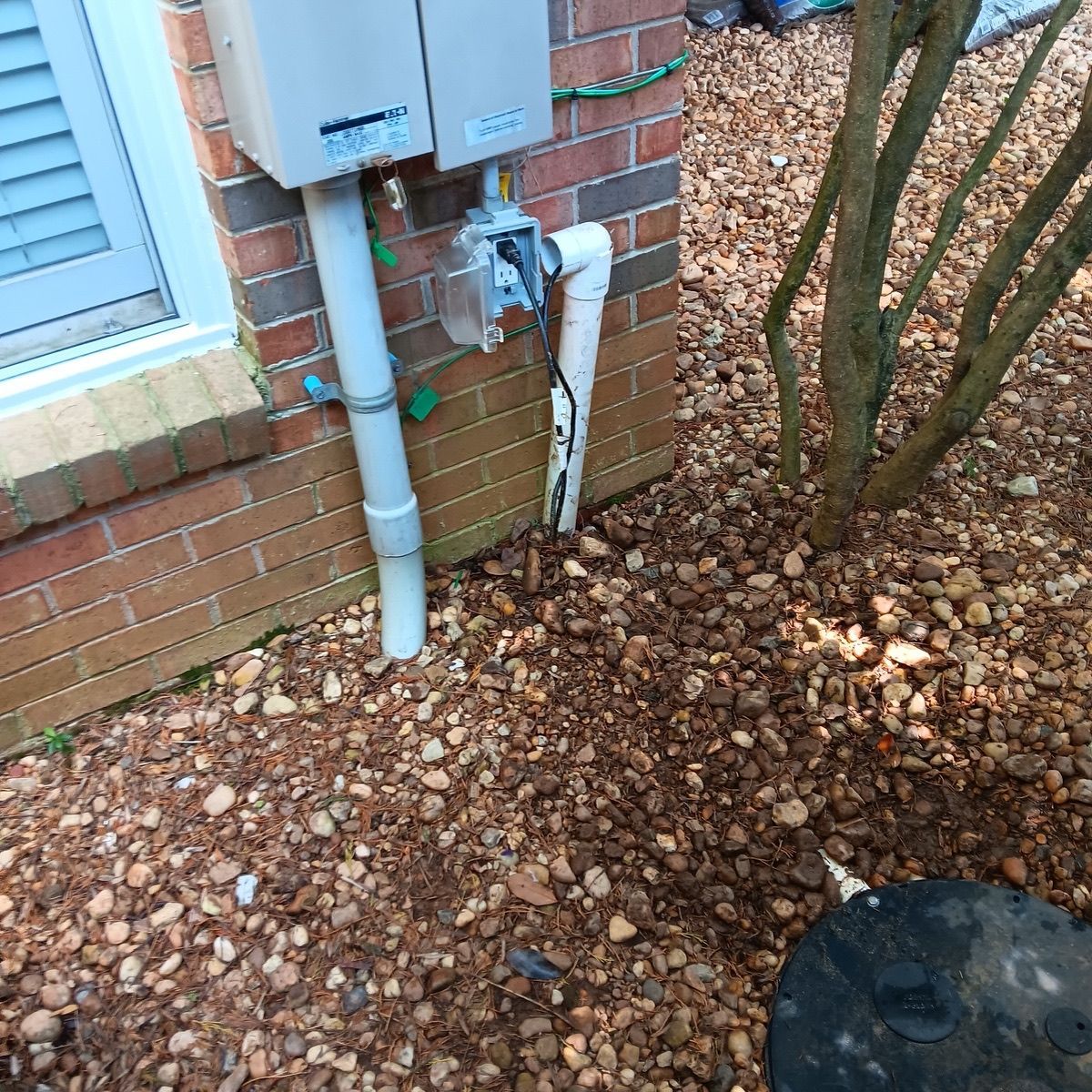 Exterior view of brick building with plumbing pipes and gas meter near gravel and a tree.