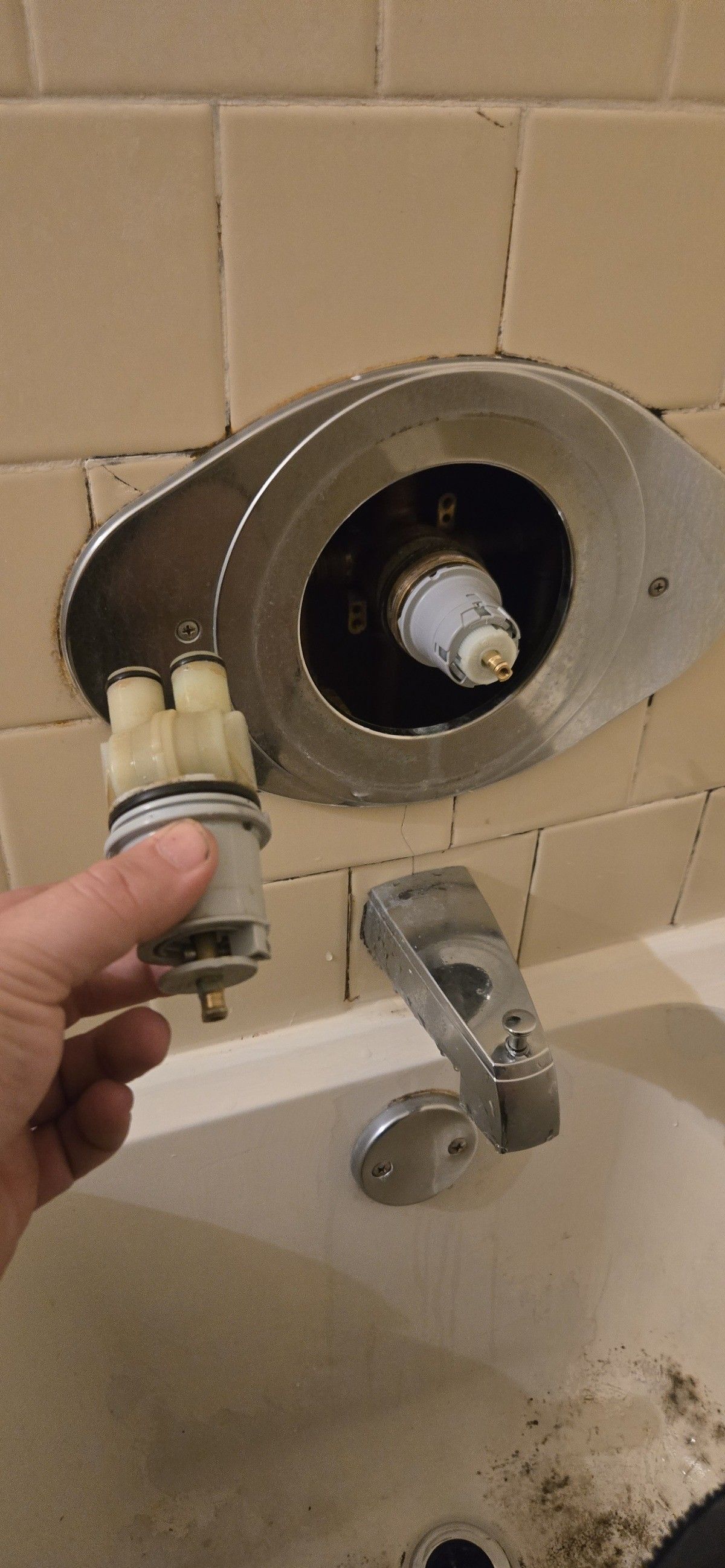 Hand holding shower valve cartridge next to a shower valve body in a tiled bathroom.