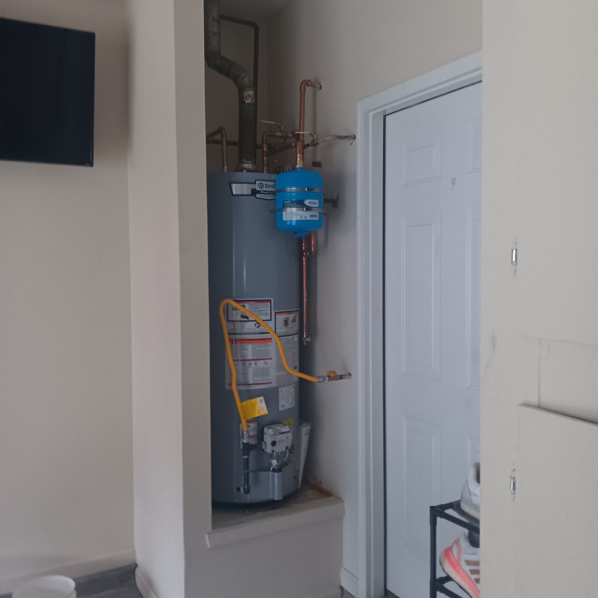 A water heater with copper pipes in a recessed alcove, next to a white door. A blue expansion tank is on top.