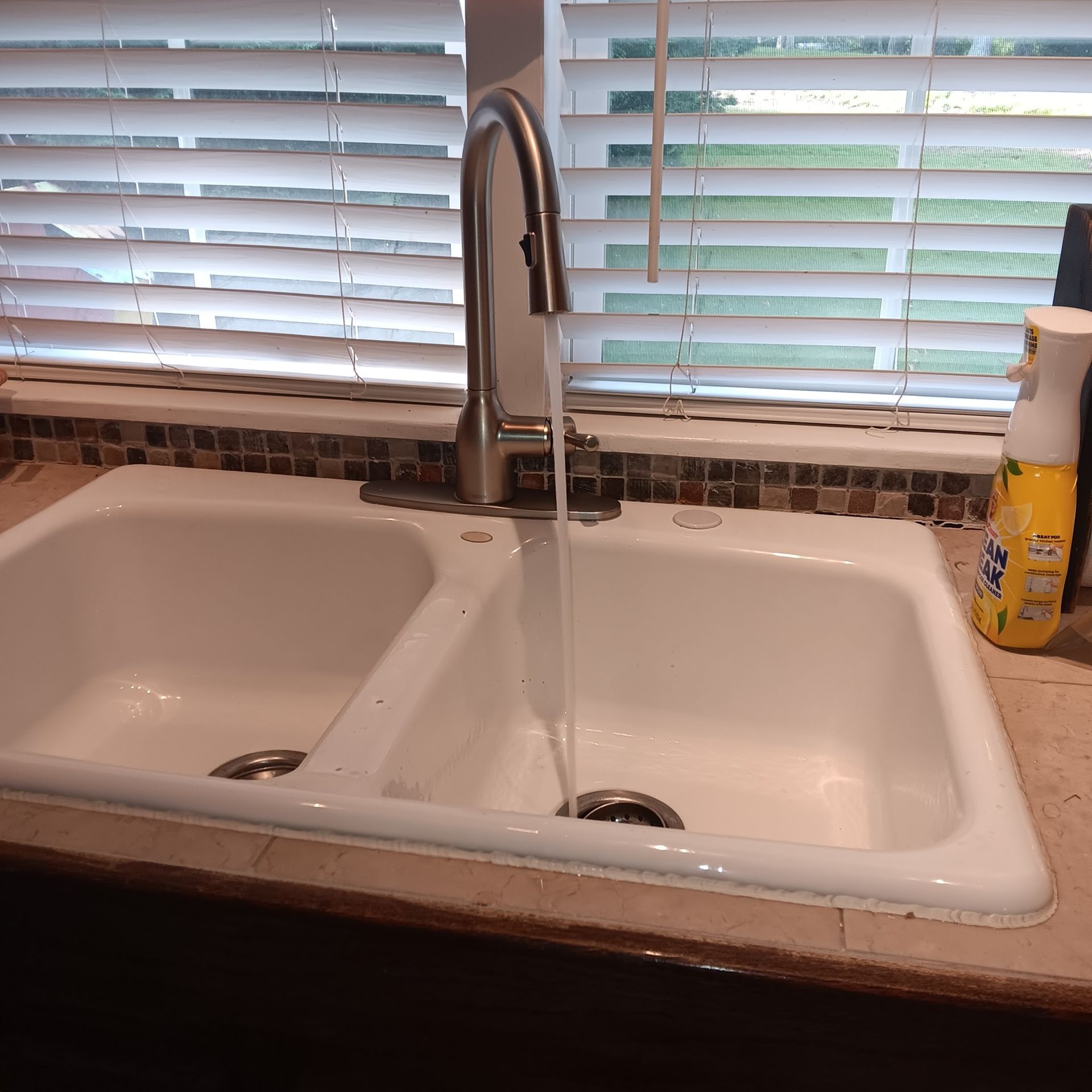 Kitchen sink with running water, white basin, stainless steel faucet, window with blinds in the background.